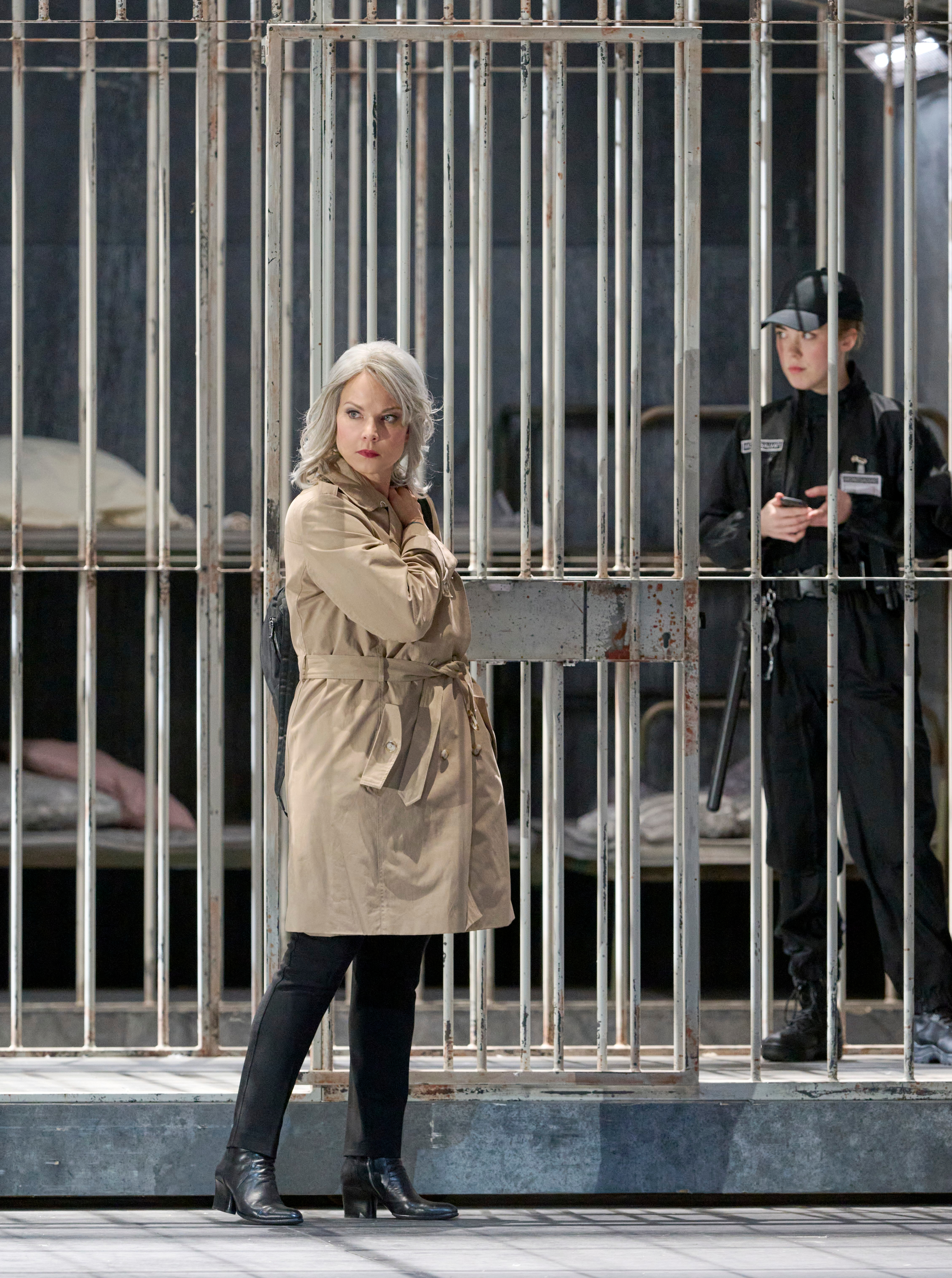 Eine Frau im Trenchcoat steht vor einem vergitterten Raum, daneben eine uniformierte Wache mit Notizblock.