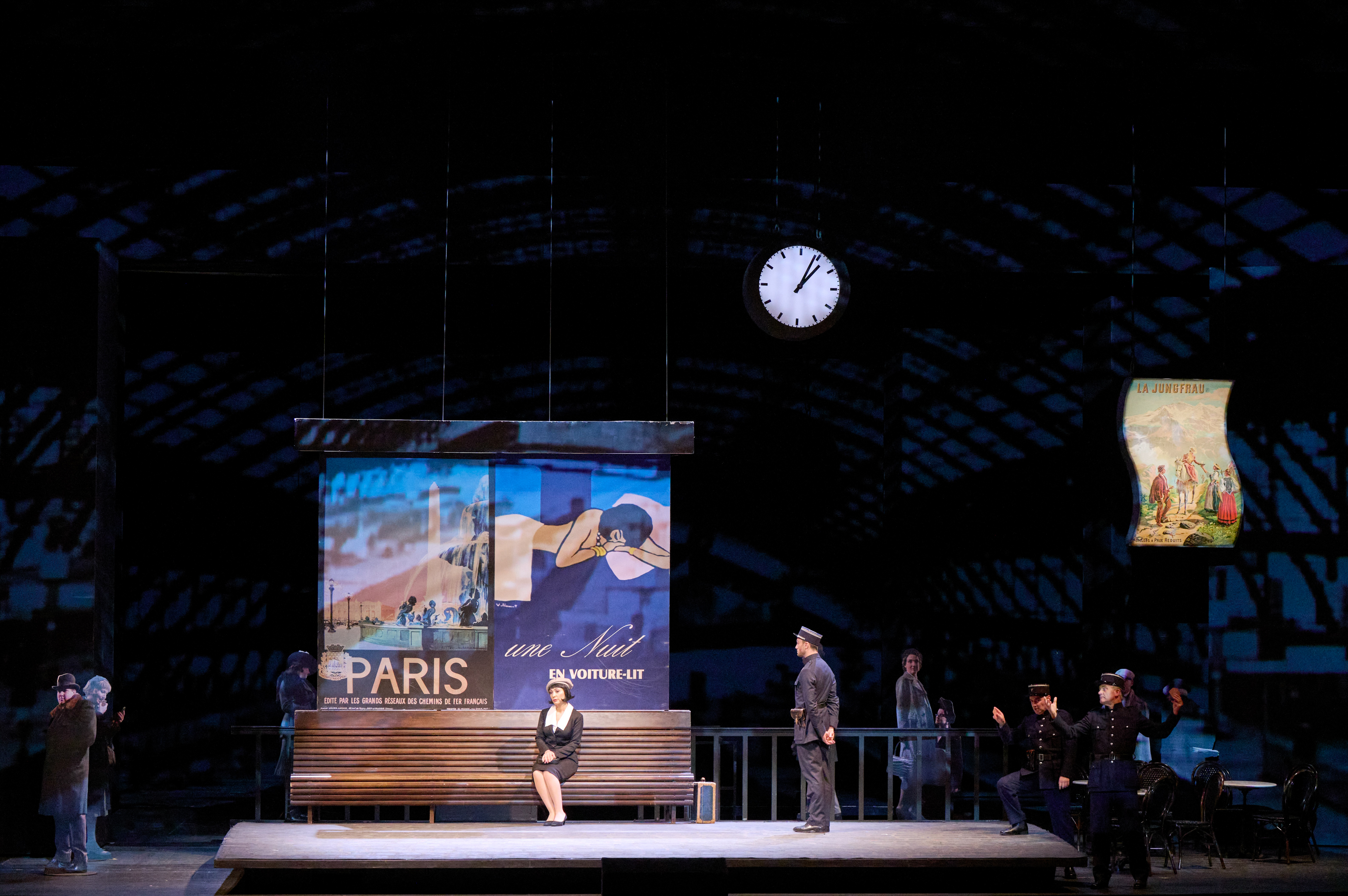 Eine Frau sitzt allein auf einer Bank unter einer Bahnhofsuhr, umgeben von Reisenden und Werbeplakaten auf einer Theaterbühne.