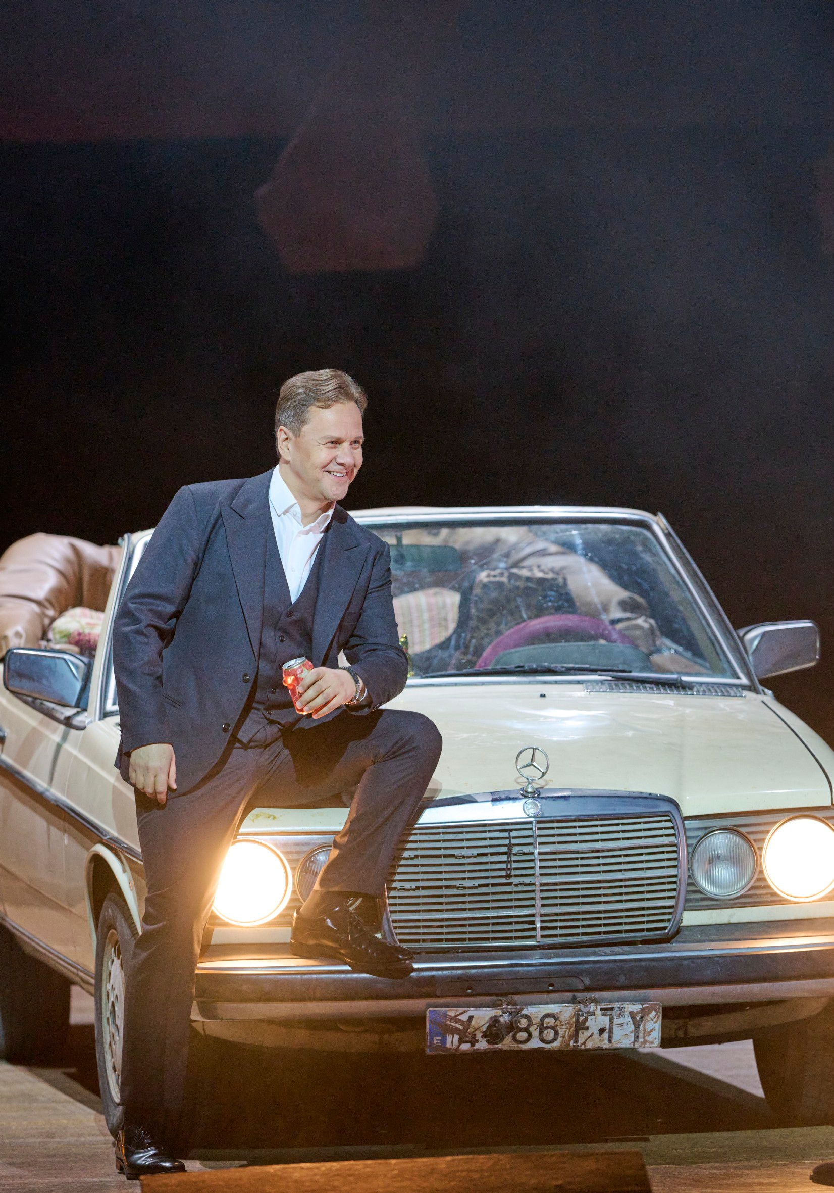 Ein Mann im Anzug lehnt lässig mit einer Getränke-Dose an einem weißen Oldtimer, während aus dem Autoinneren Arme und Beine herausragen.