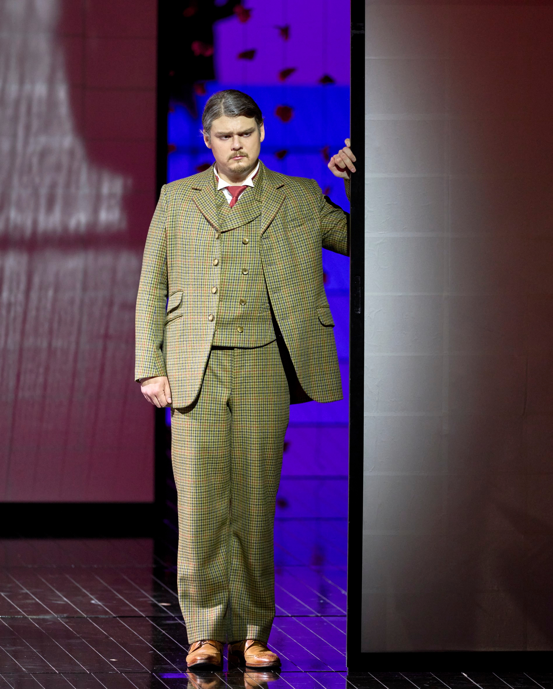 Ein Mann im braunen Anzug steht nachdenklich in einem Türrahmen vor farbig beleuchtetem Bühnenhintergrund.