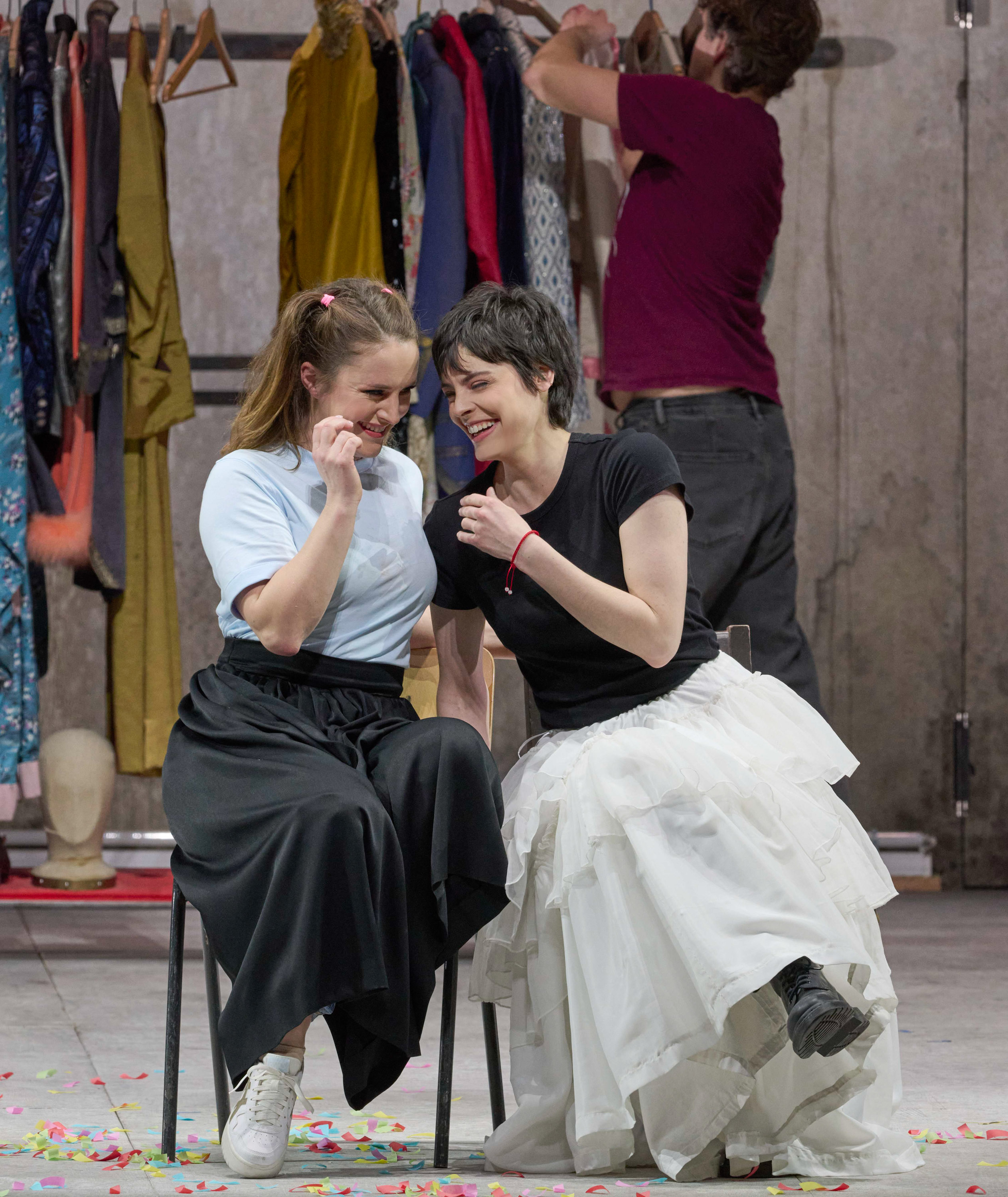 Zwei Frauen sitzen lachend nebeneinander vor einer Kleiderstange mit bunten Kostümen; im Hintergrund hängt ein Mann neue Kleidung auf.