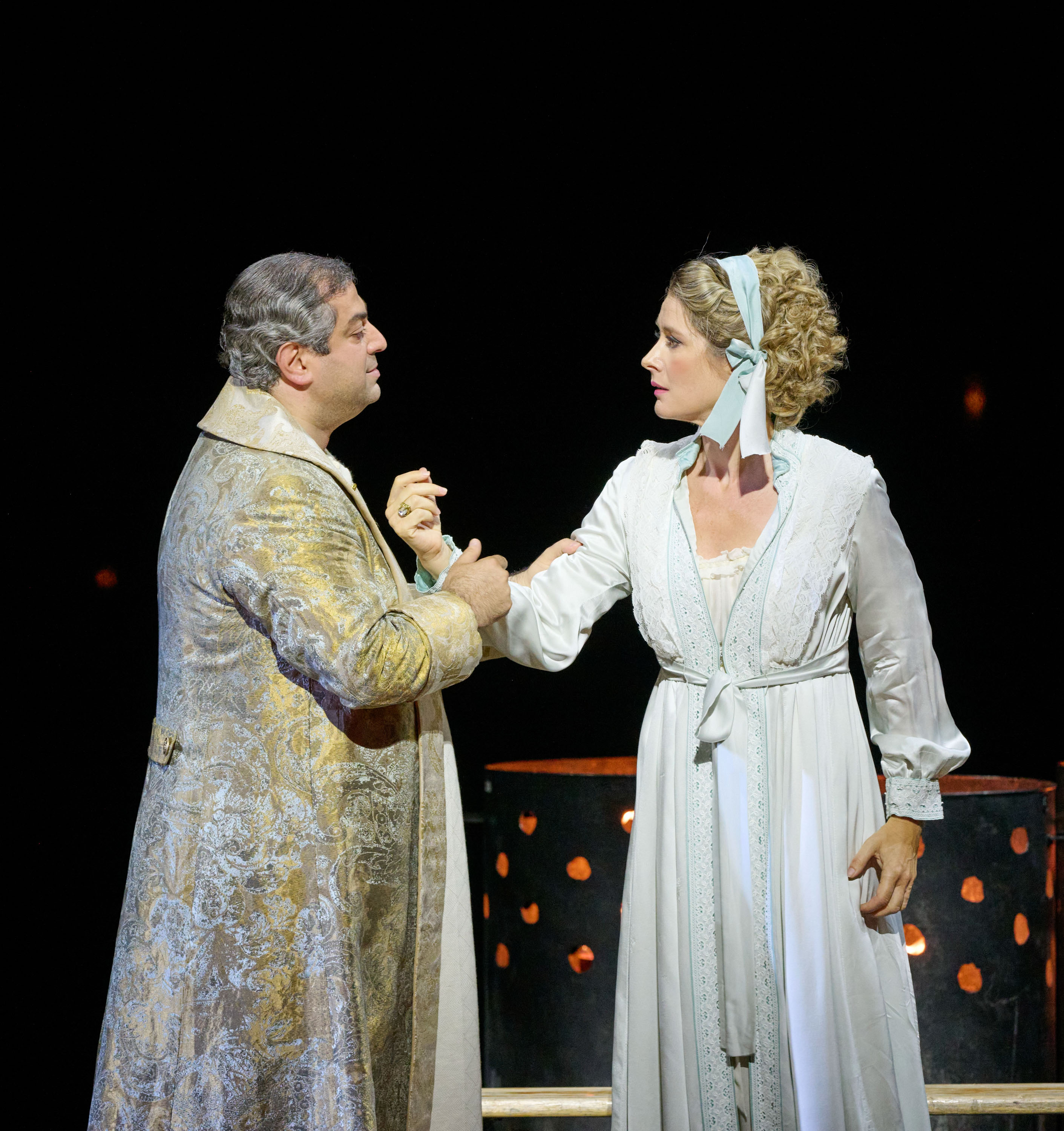 Eine Frau in einem weißen Kleid steht einem Mann in einem goldenen Morgenmantel gegenüber. Er hält sie am Arm fest.