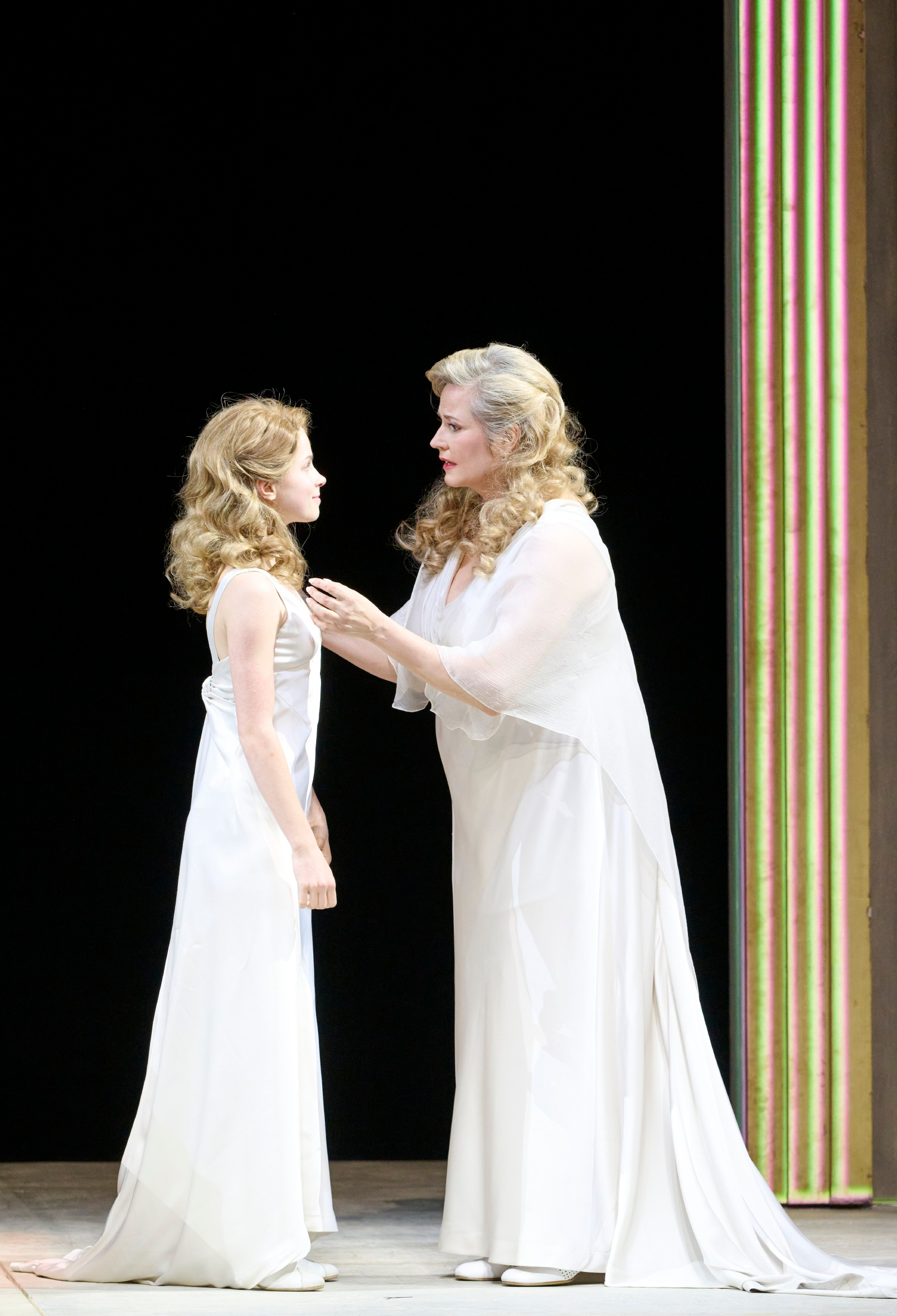 Zwei Frauen in weißen Kleidern stehen sich vor einem schwarzen Hintergrund gegenüber, eine legt der anderen beruhigend die Hände auf die Schultern.