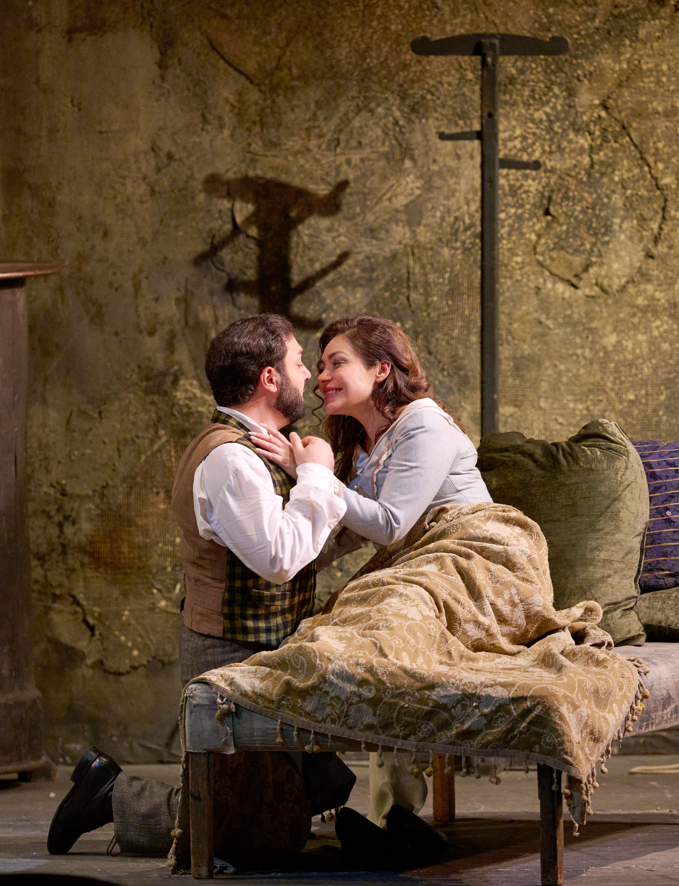 Ein Mann kniet vor einem Bett, während er liebevoll das Gesicht einer Frau berührt, die unter einer Decke sitzt und ihn anlächelt.