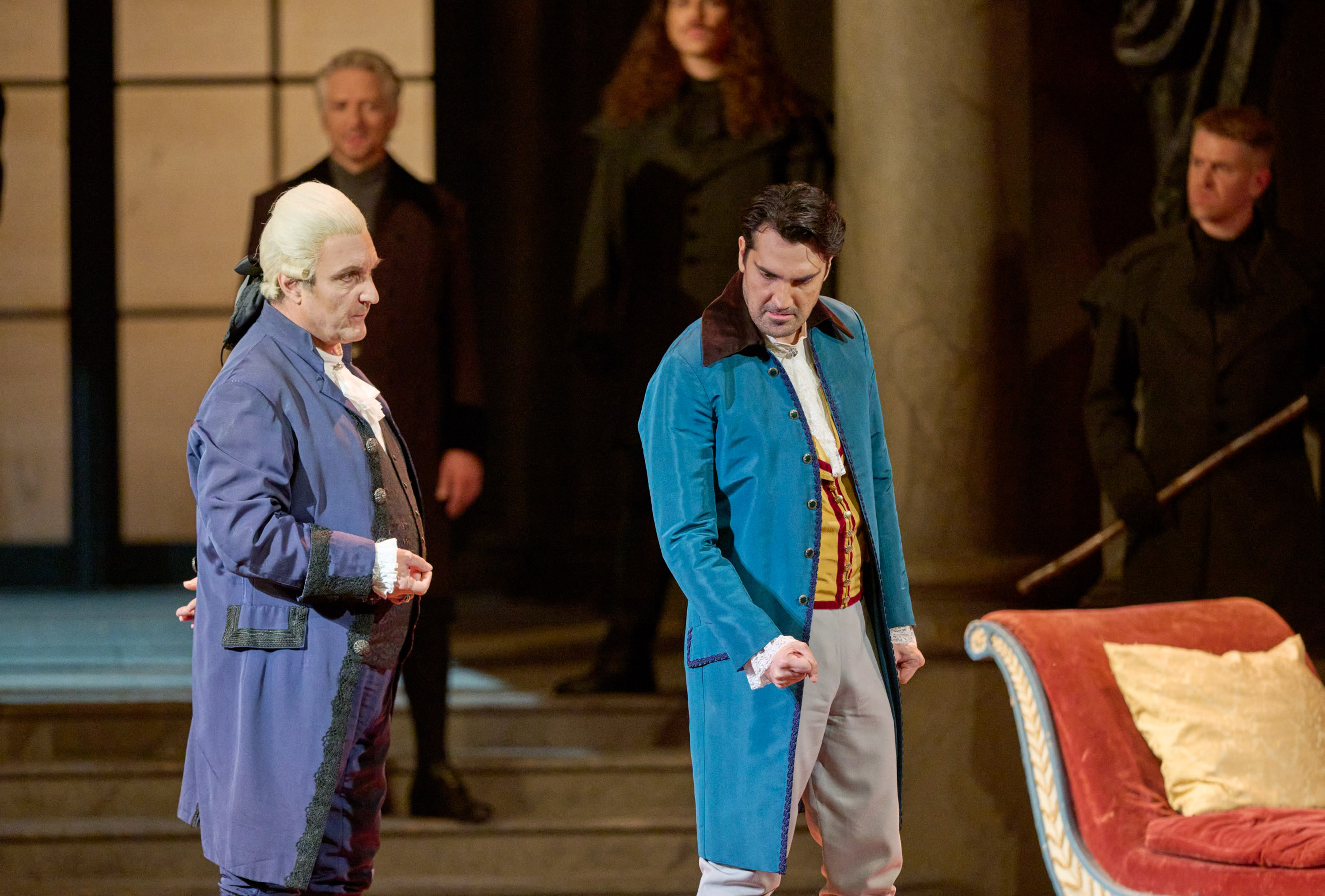 Ein Mann in einem blauen Kostüm steht mit geballter Faust in der rechten Hälfte des Bildes. Hinter ihm steht ein Mann, der eine weiße Perücke und ein violettes Kostüm trägt.