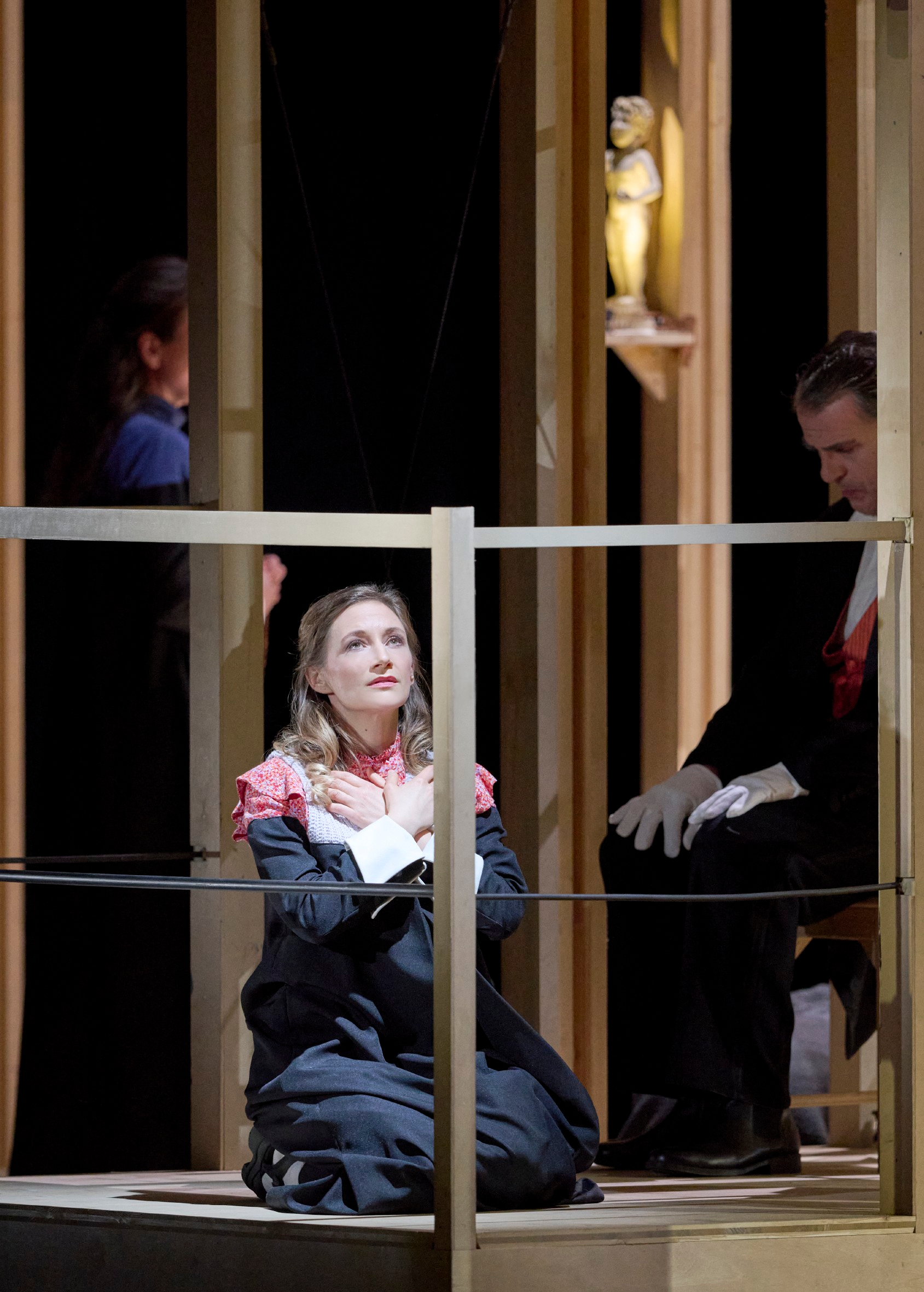 Eine Frau in dunkler Kleidung kniet mit verschränkten Armen und bittendem Blick, während ein Mann in Frack neben ihr sitzt und eine weitere Person im Hintergrund steht.