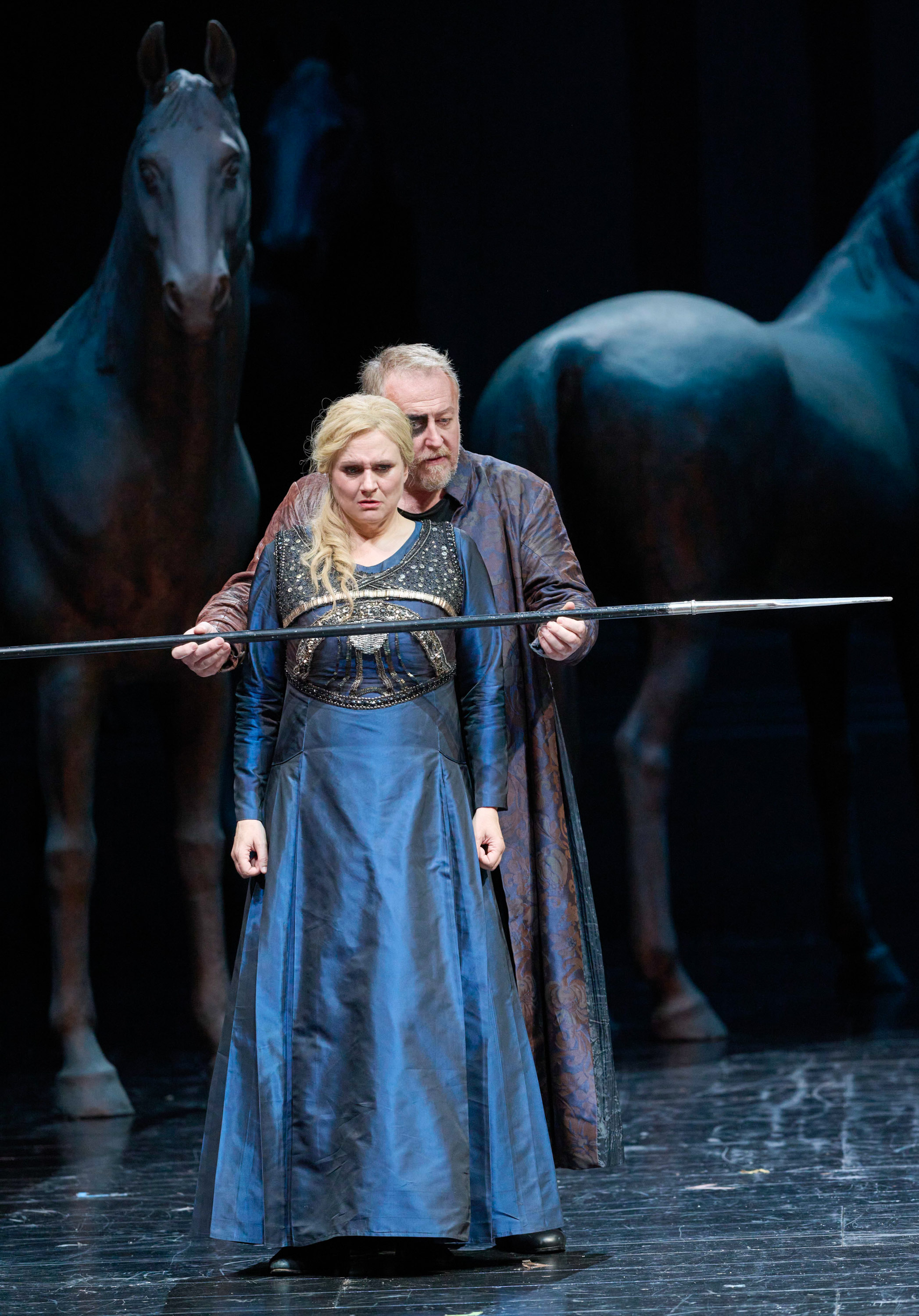 Eine blonde Frau in einem langen blauen Kleid wird von einem Mann in einem langen Mantel von hinten umschlungen, während er einen Stab vor ihr hält. 