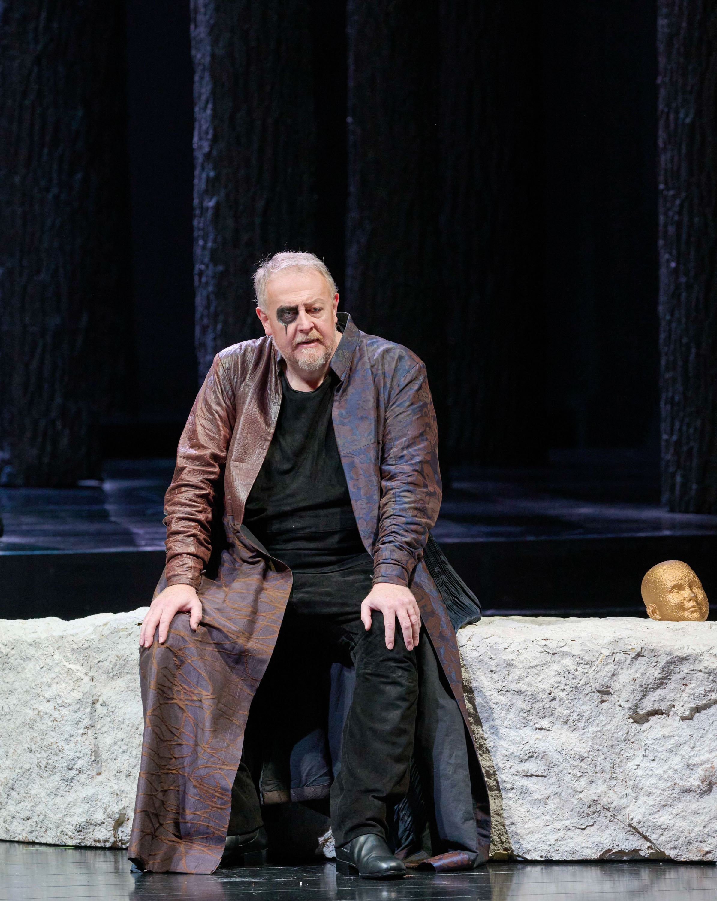 Ein Mann in langem Ledermantel sitzt auf einer Steinmauer mit einer Skulptur eines Kopfes daneben.