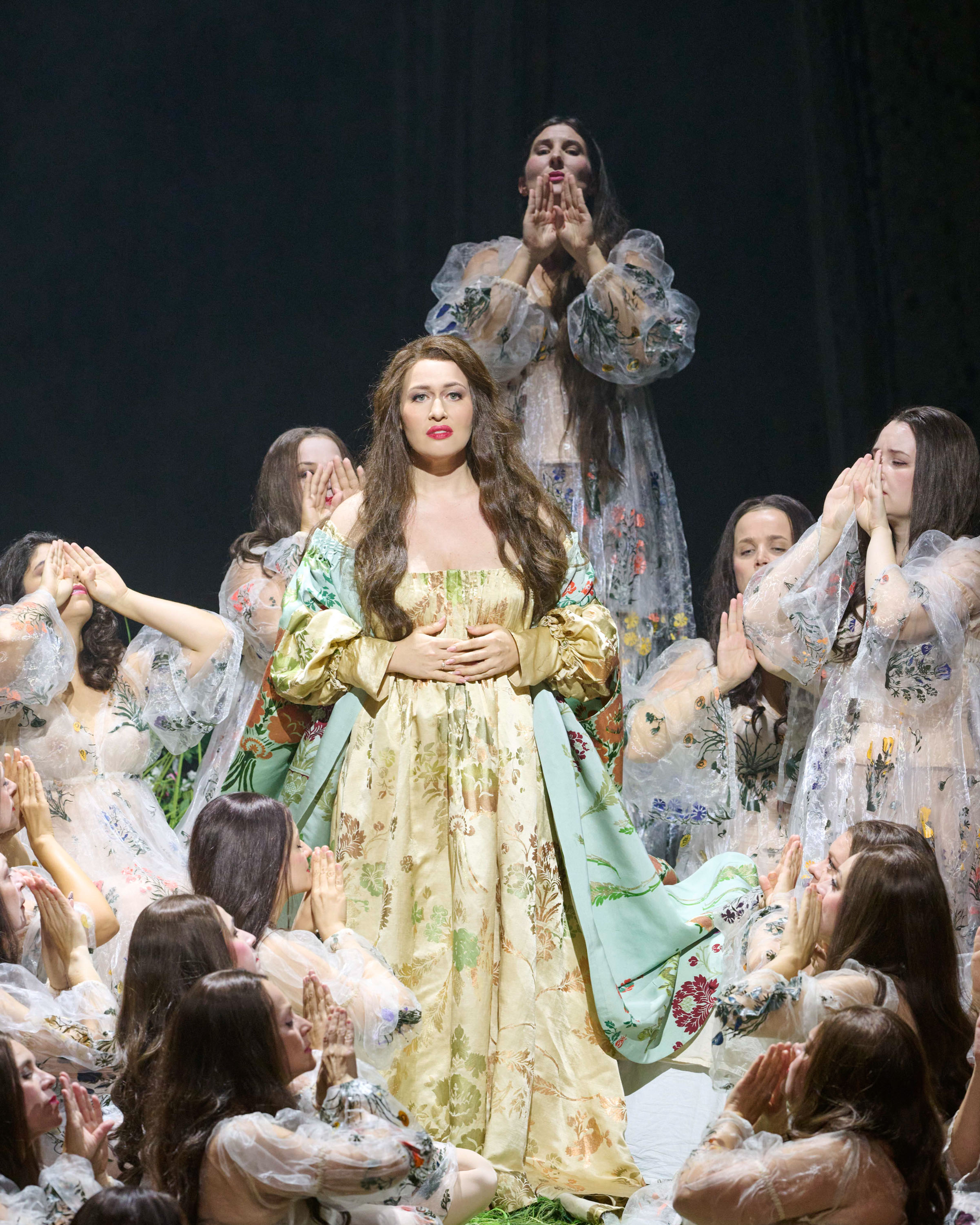 Eine langhaarige Frau steht in einem langen hellgelben Gewand in der Mitte. Um sie herum stehen weitere Frauen, die ihr etwas zurufen.