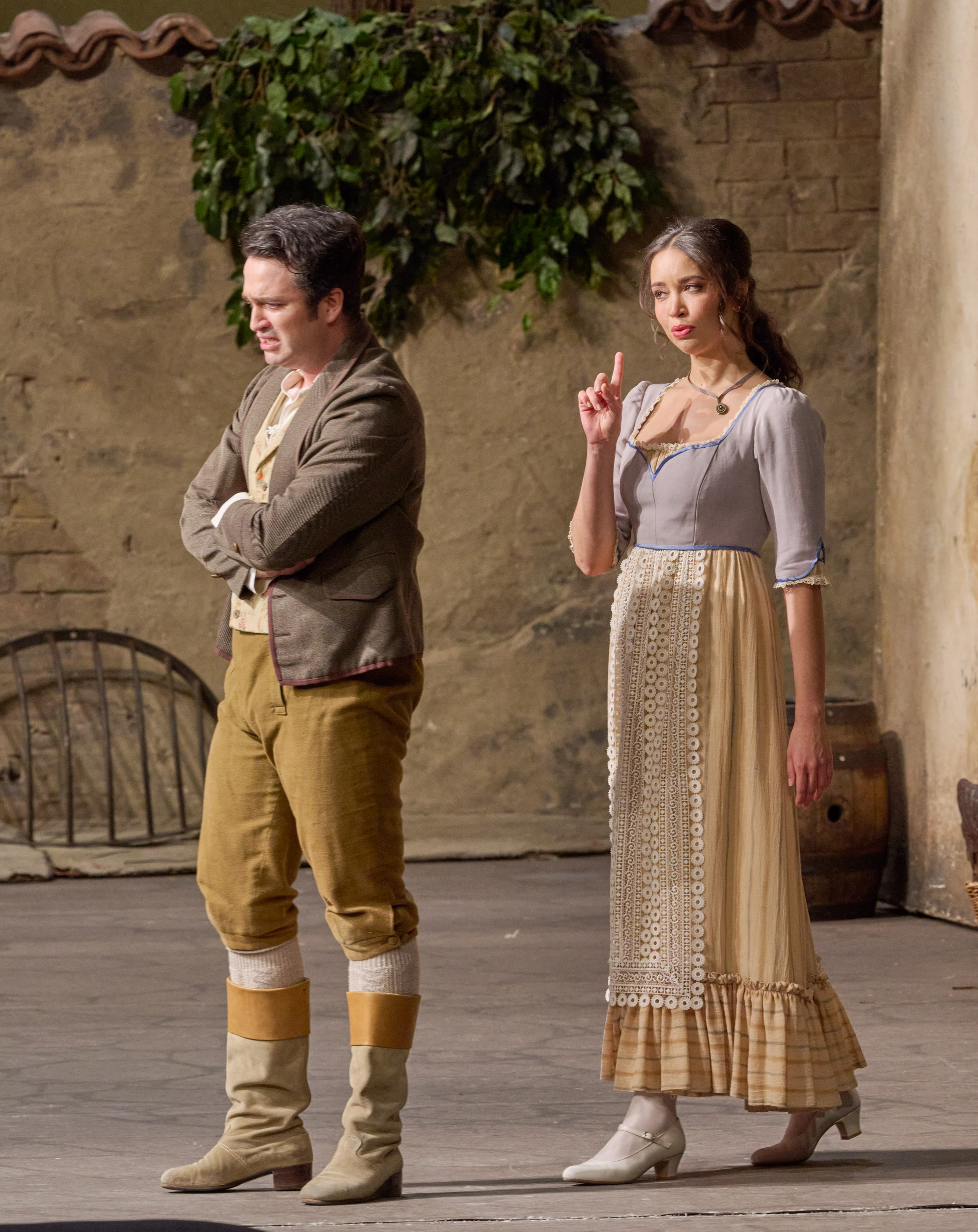 Eine Frau hebt mahnend den Finger, während ein Mann neben ihr mit verschränkten Armen abgewandt steht, beide in historischer Kleidung auf einer Bühne.