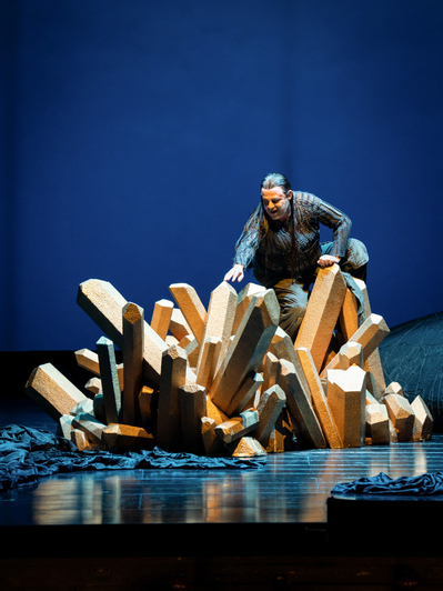 Eine Figur klettert aus einer Ansammlung goldfarbener Kristallformen auf einer Bühne mit dunklem Hintergrund.