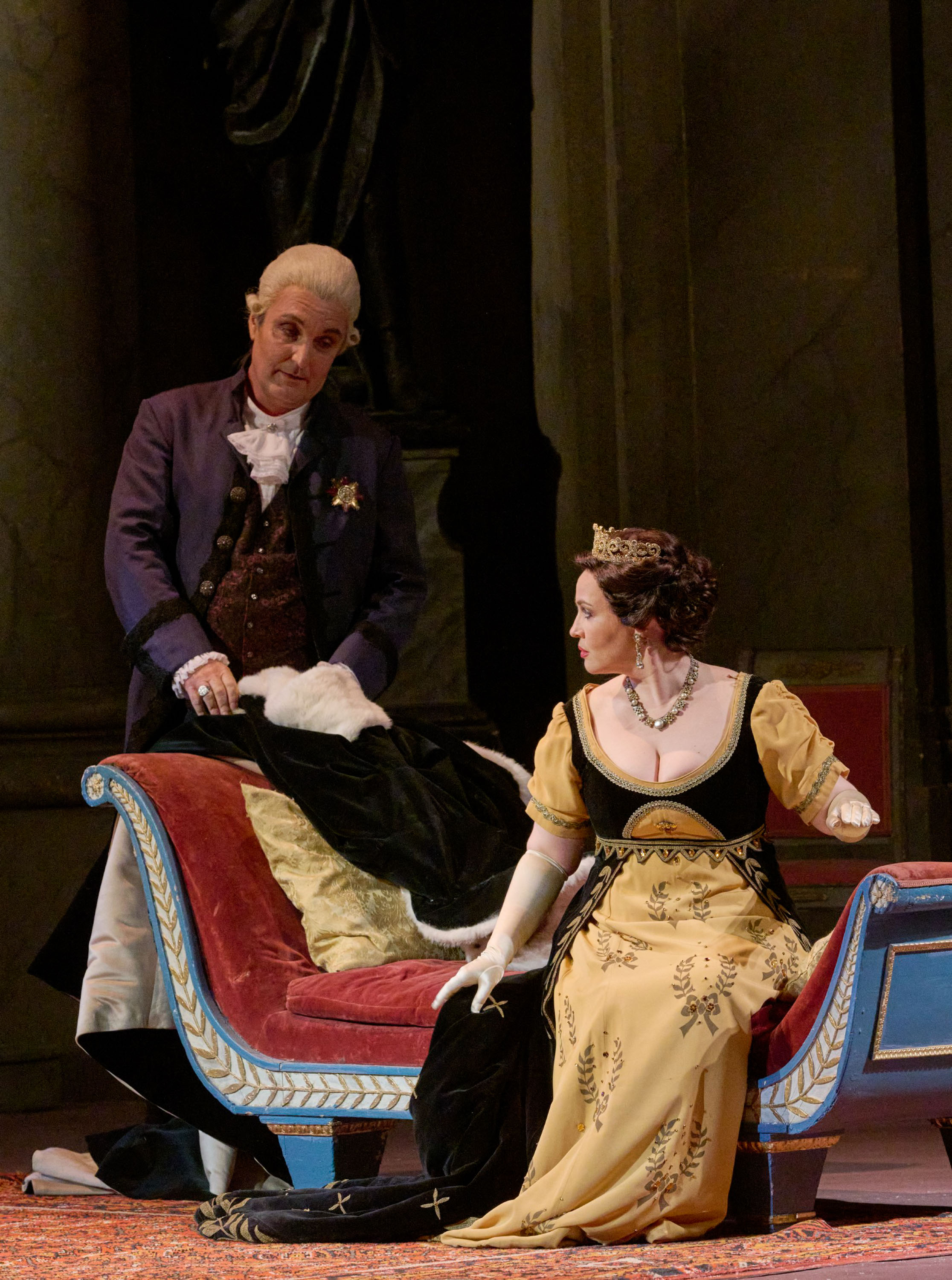 Eine Dame in einem schwarz-gelben Kleid und einer Krone auf dem Kopf sitzt auf einem eher prunkvoll aussehendem Sitzmöbel. Dahinter steht ein Mann mit einer weißen Perücke auf dem Kopf. Sie dreht sich zu ihm um. 