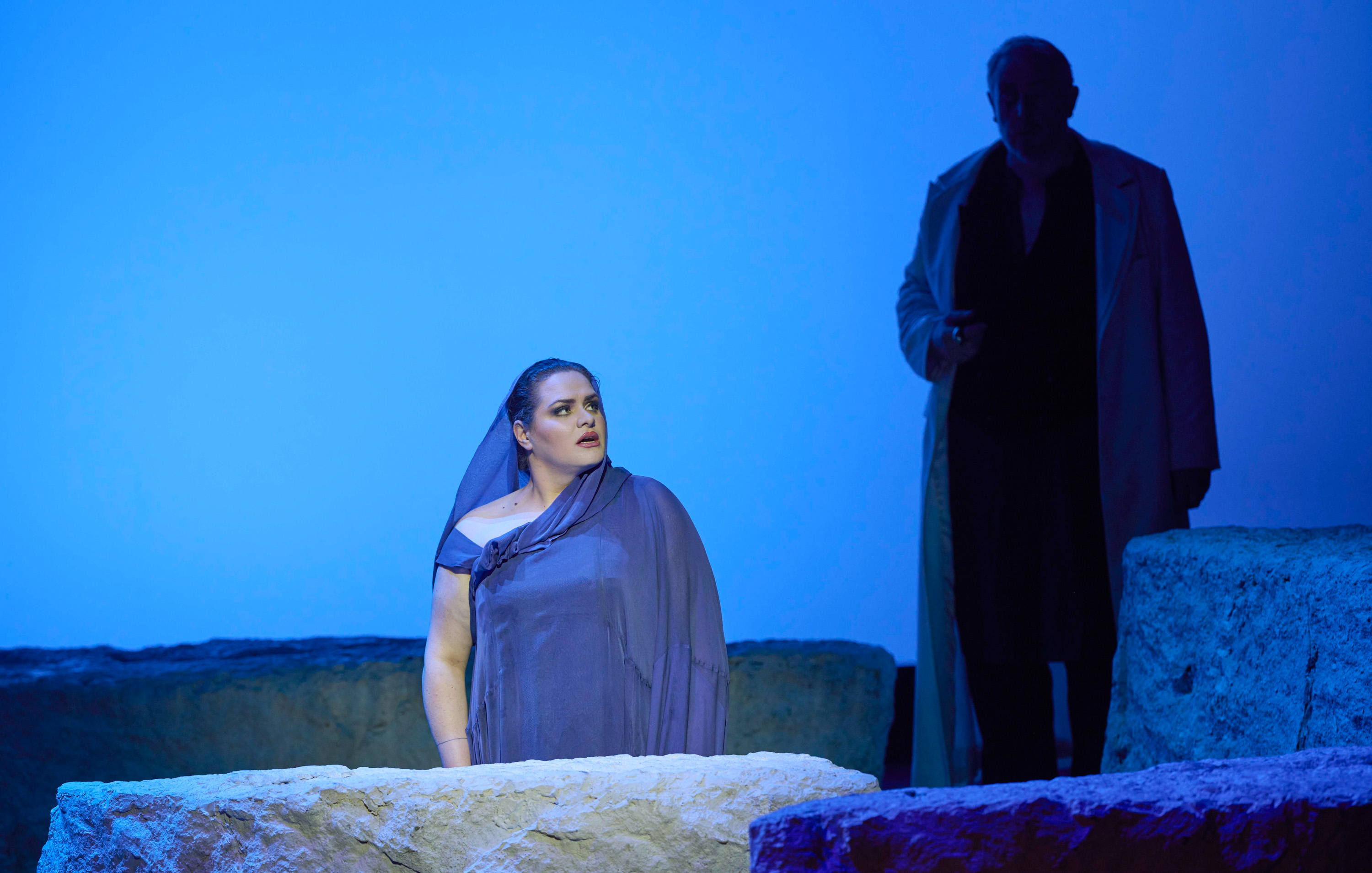 Eine Frau in einem lila Gewand schaut ernst in die Ferne, während ein Mann im Hintergrund steht.