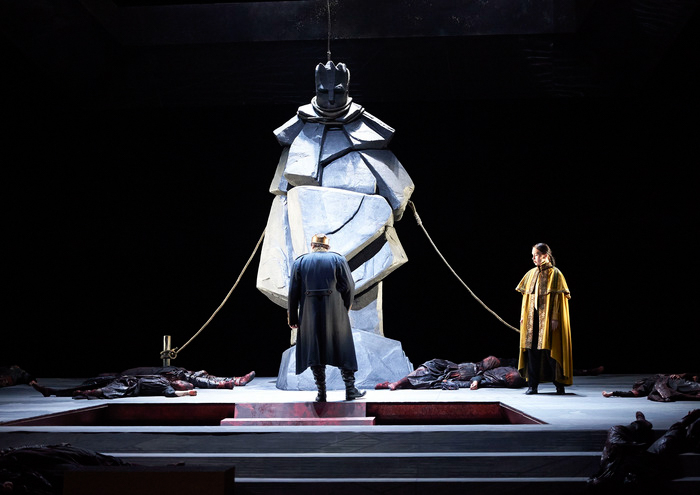Eine imposante Statue steht im Zentrum der Bühne, umgeben von am Boden liegenden Figuren, während zwei Männer in dunklen Mänteln davor stehen.