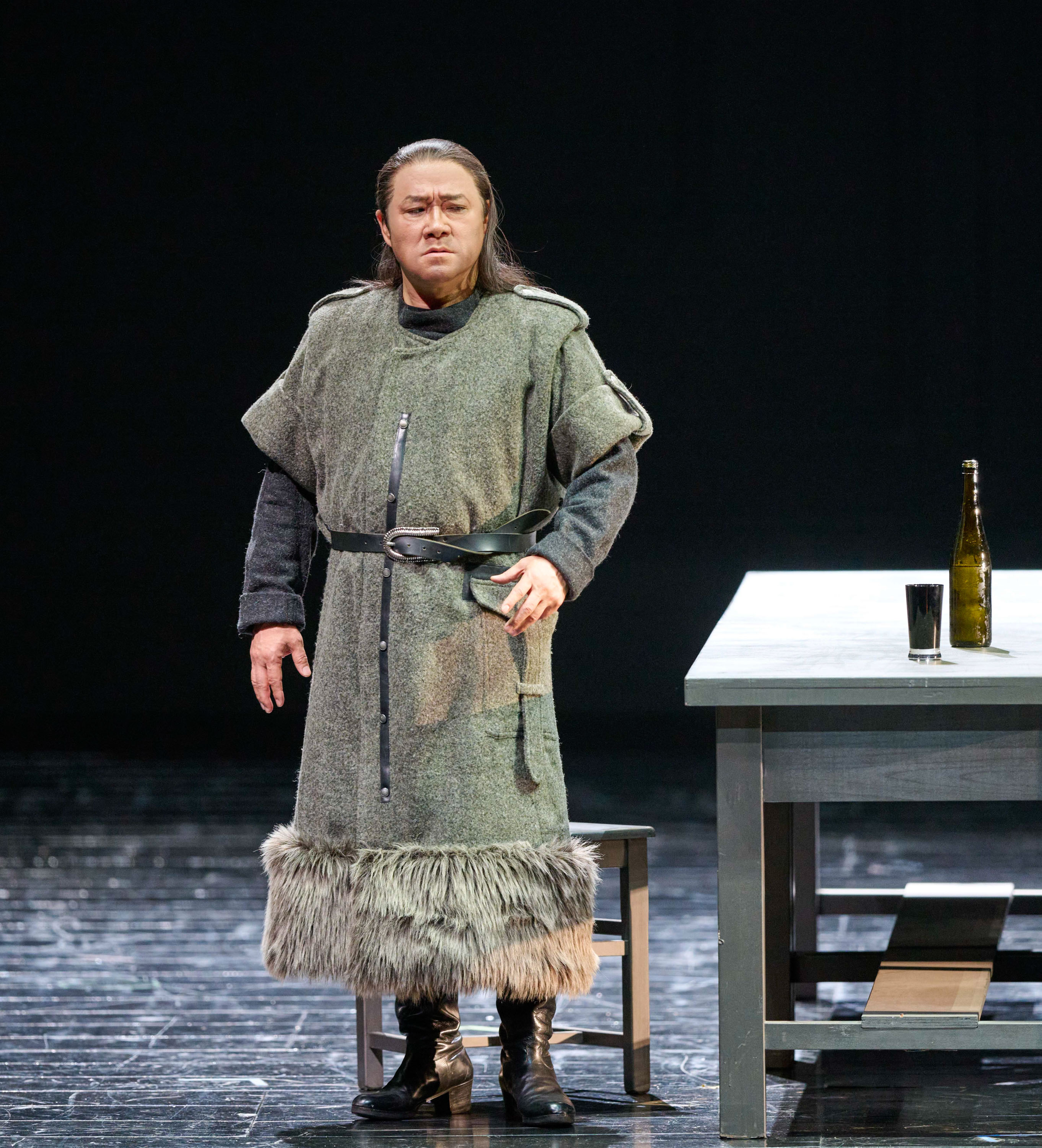 Ein Mann in mittelalterlicher Gewandung steht auf einer Bühne neben einem Tisch mit einer Flasche und einem Becher.