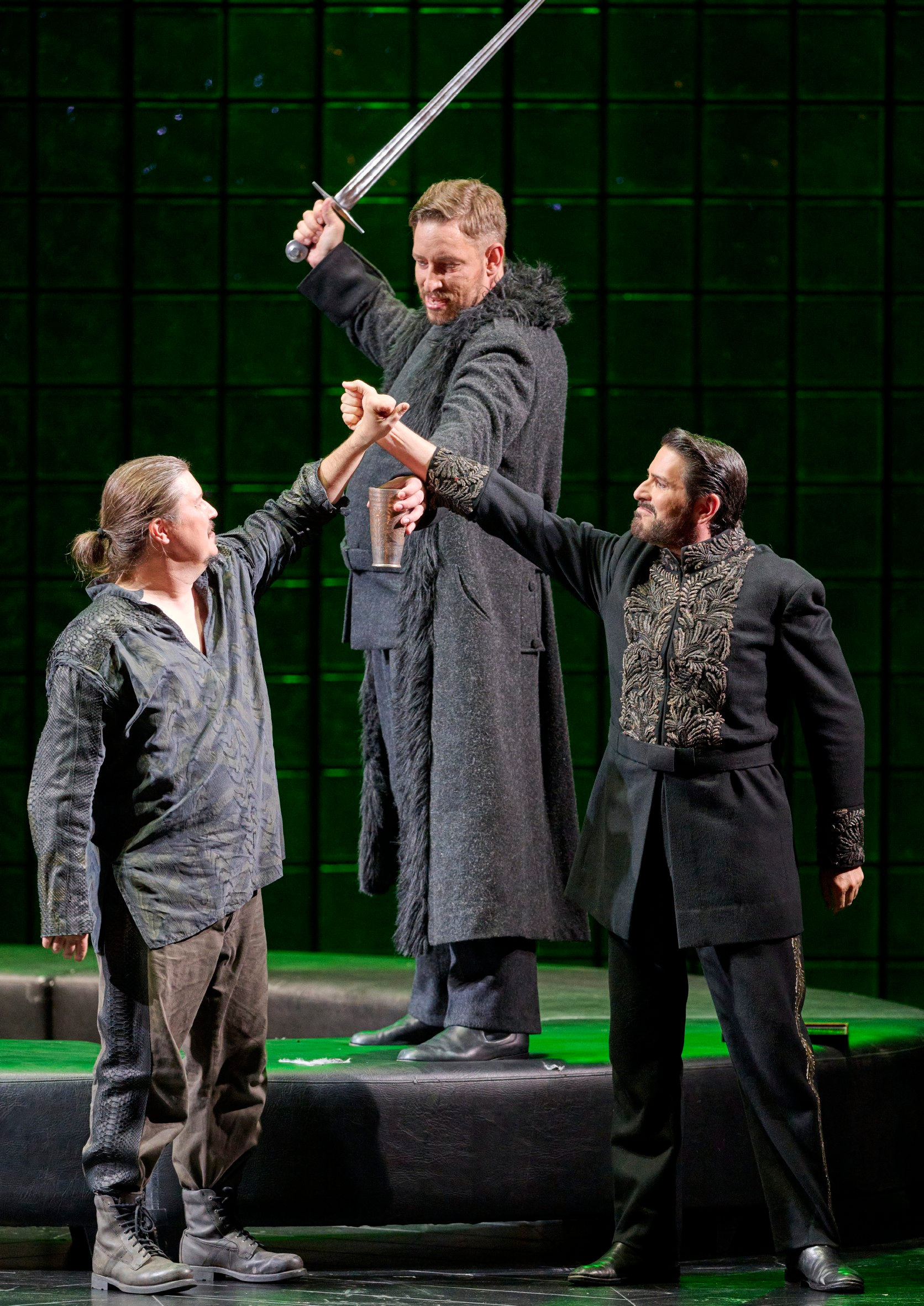 Drei Männer stehen auf der Bühne vor grünem Hintergrund, während zwei von ihnen den Arm des Dritten festhalten, der ein Schwert in die Höhe hebt.