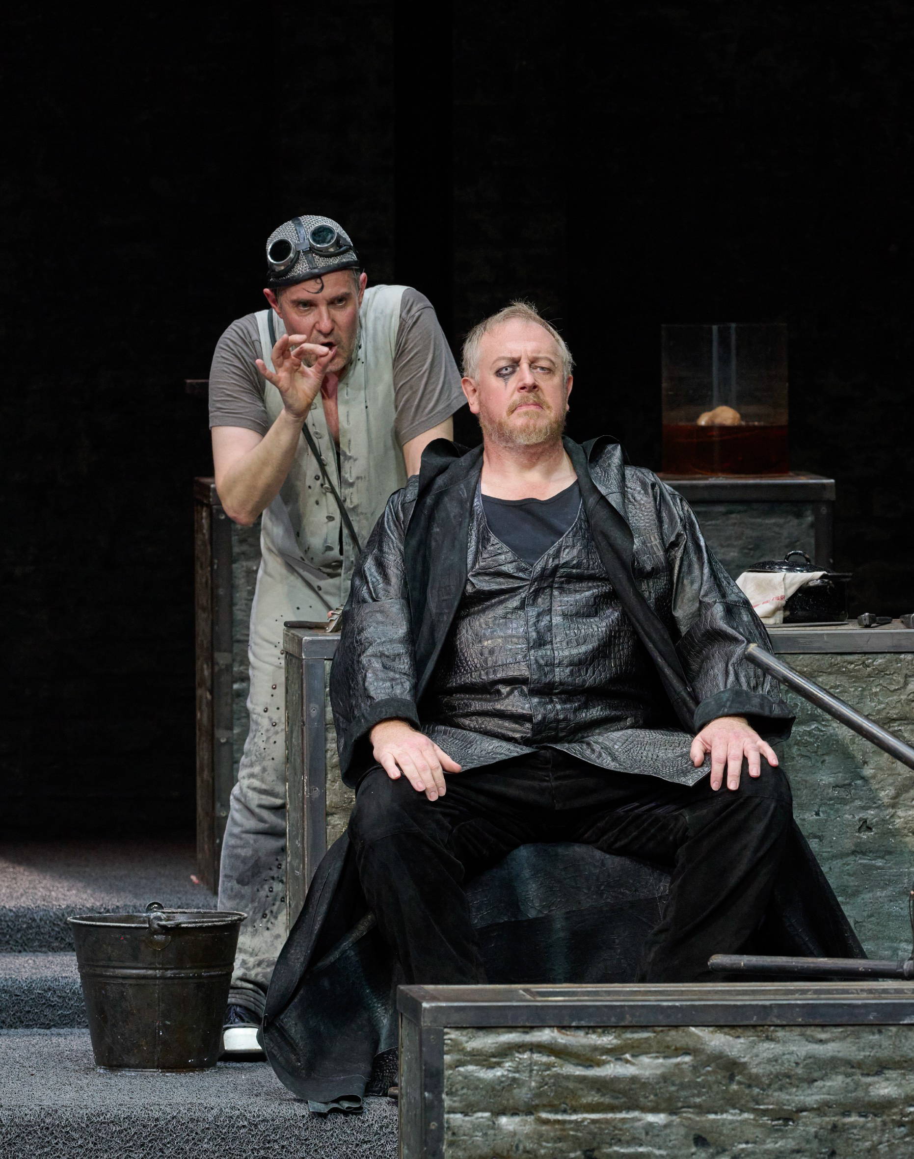 Zwei Männer vor dunklem Hintergrund; der eine sitzt, der andere steht dahinter und beide blicken nach vorne.