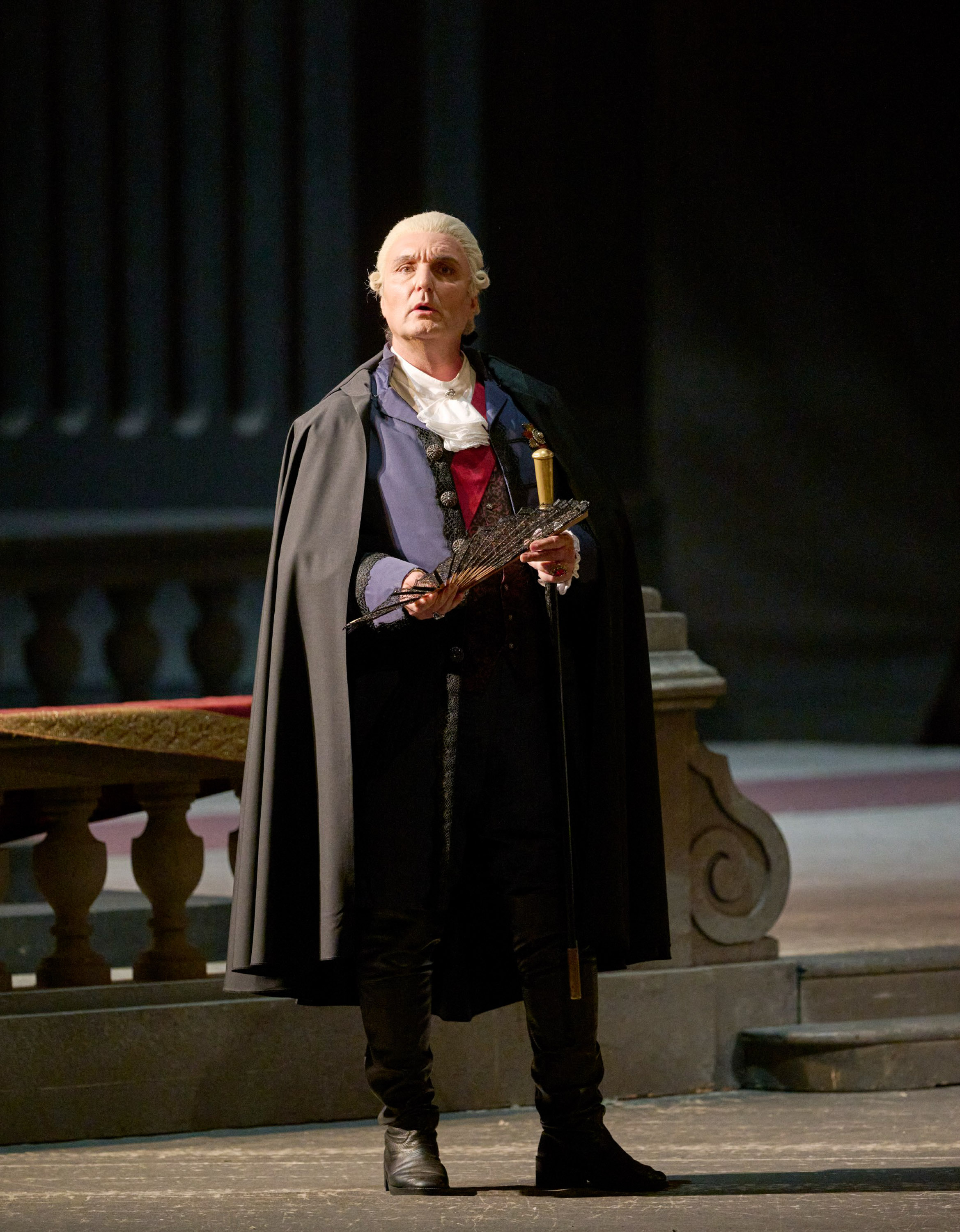 Man sieht einen Mann in einem dunklen Umhang, der vor einem dunklen Hintergrund steht. Er trägt eine weiße Perücke auf dem Kopf und hält einen Fächer in der Hand.