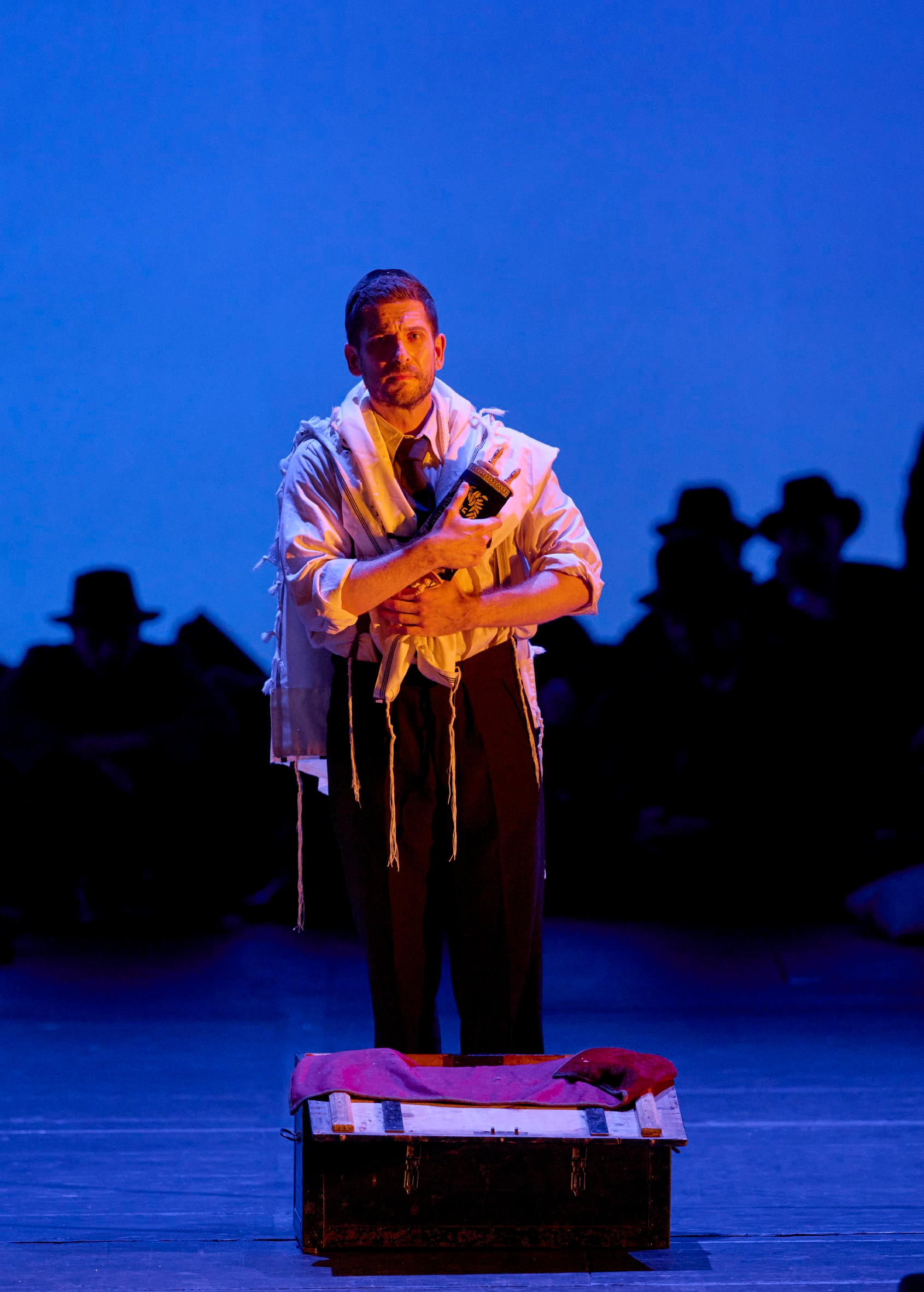 Ein Mann im Gebetsschal steht allein im Licht vor blauem Bühnenhintergrund, während im Dunkel dahinter Silhouetten mit Hüten sitzen.