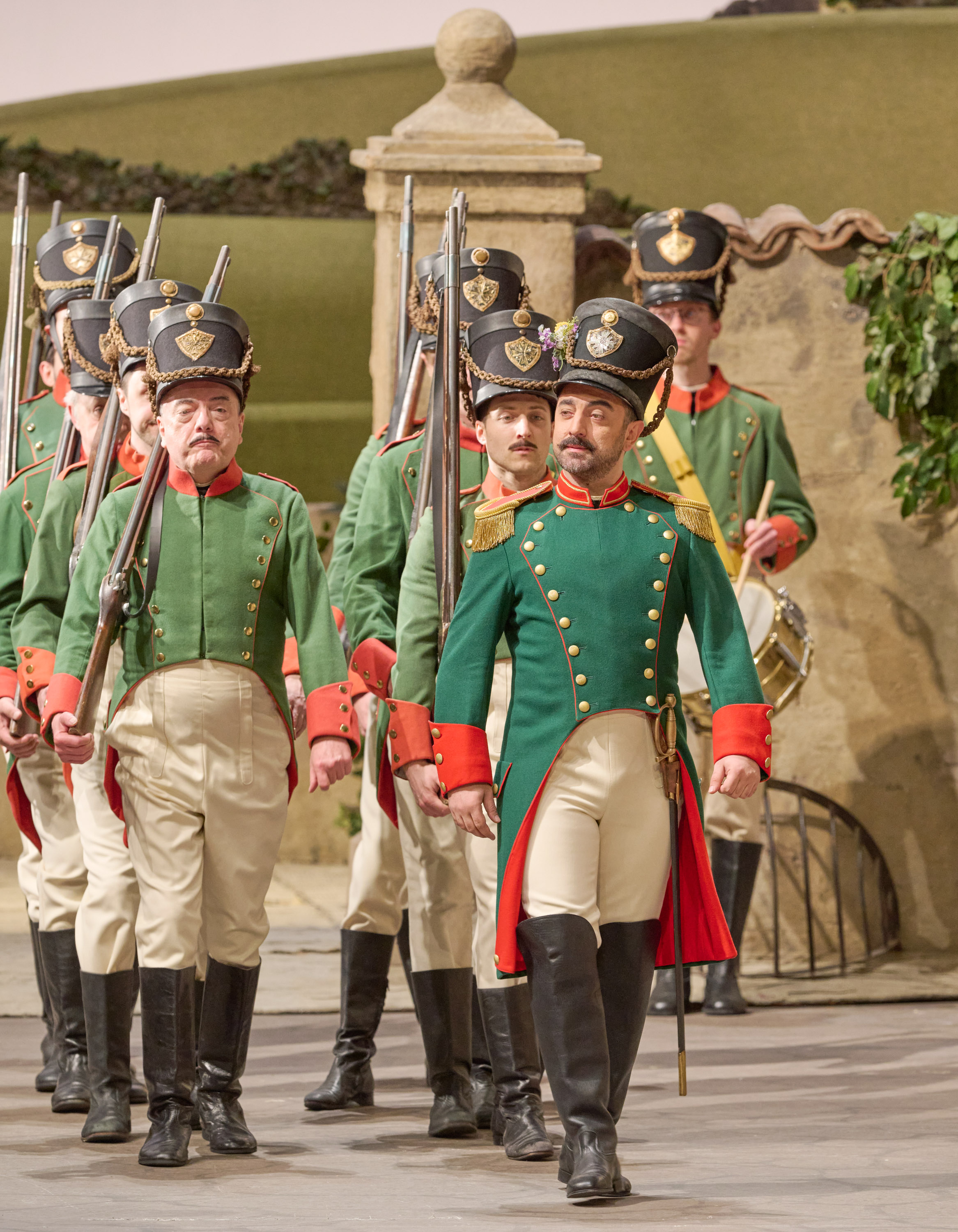 Eine Gruppe von Soldaten in grünen Uniformen marschiert mit Gewehren über eine Opernbühne im historischen Stil.