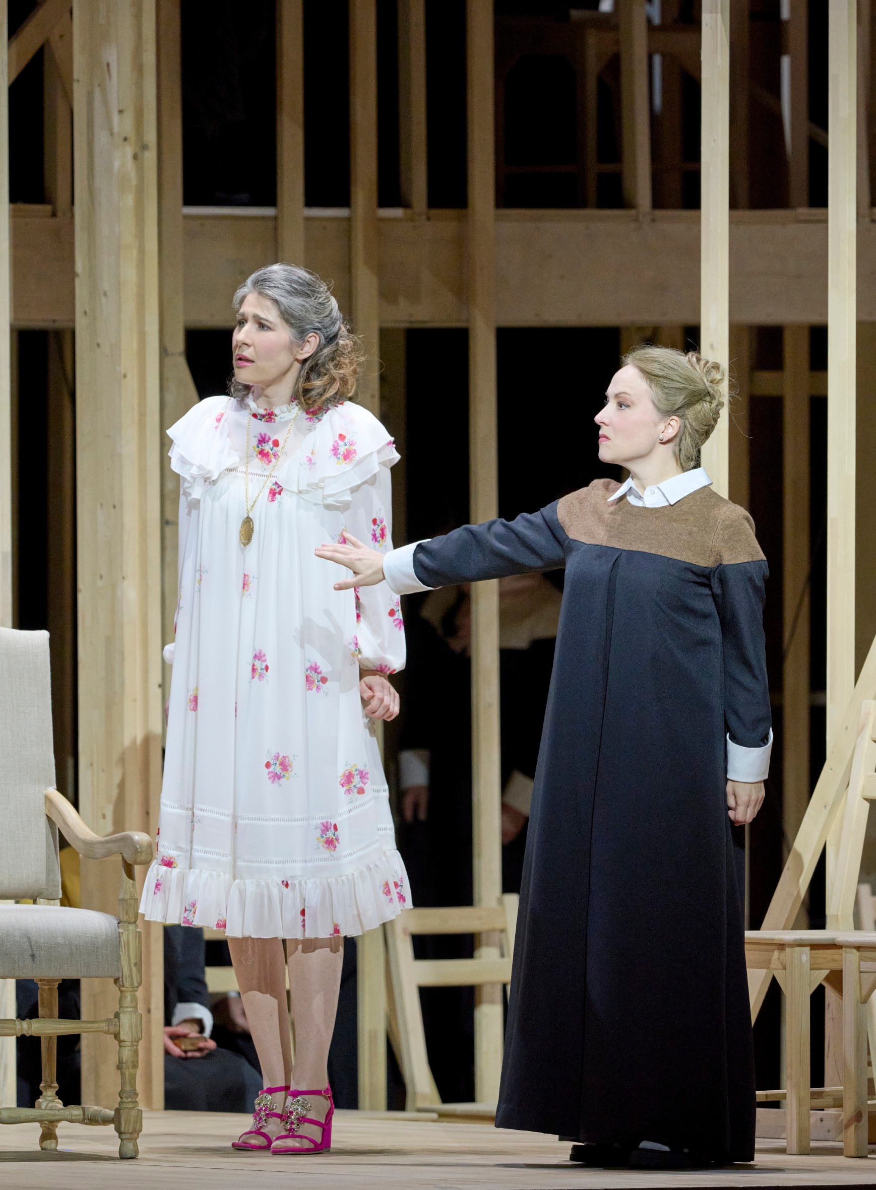 Zwei Frauen stehen vor einem hölzernen Bühnenbild; die eine trägt ein geblümtes Nachthemd, die andere streckt warnend den Arm aus.