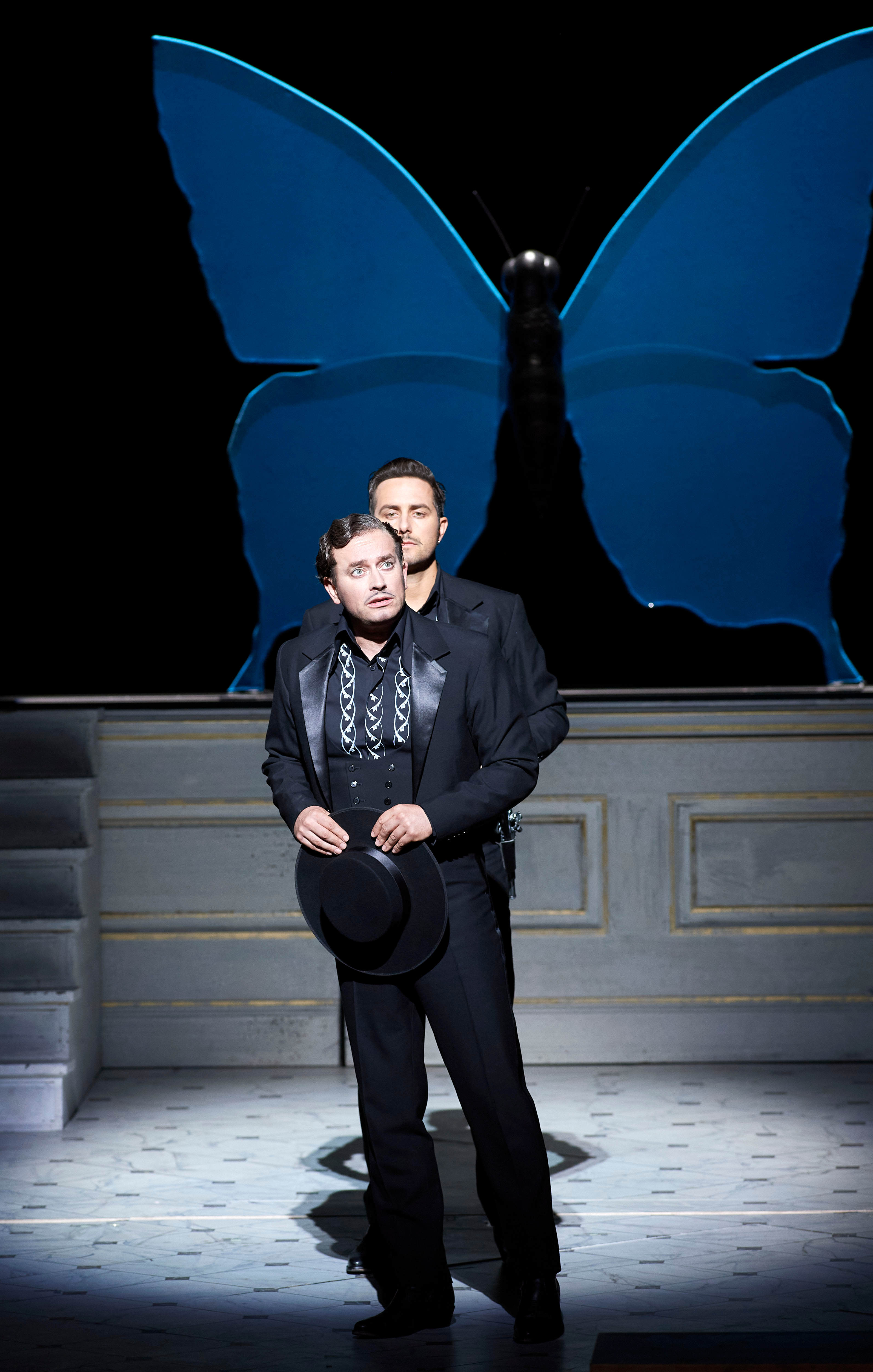 Zwei Männer in schwarzer Kleidung stehen hintereinander auf einer Bühne vor einem großen blauen Schmetterling.