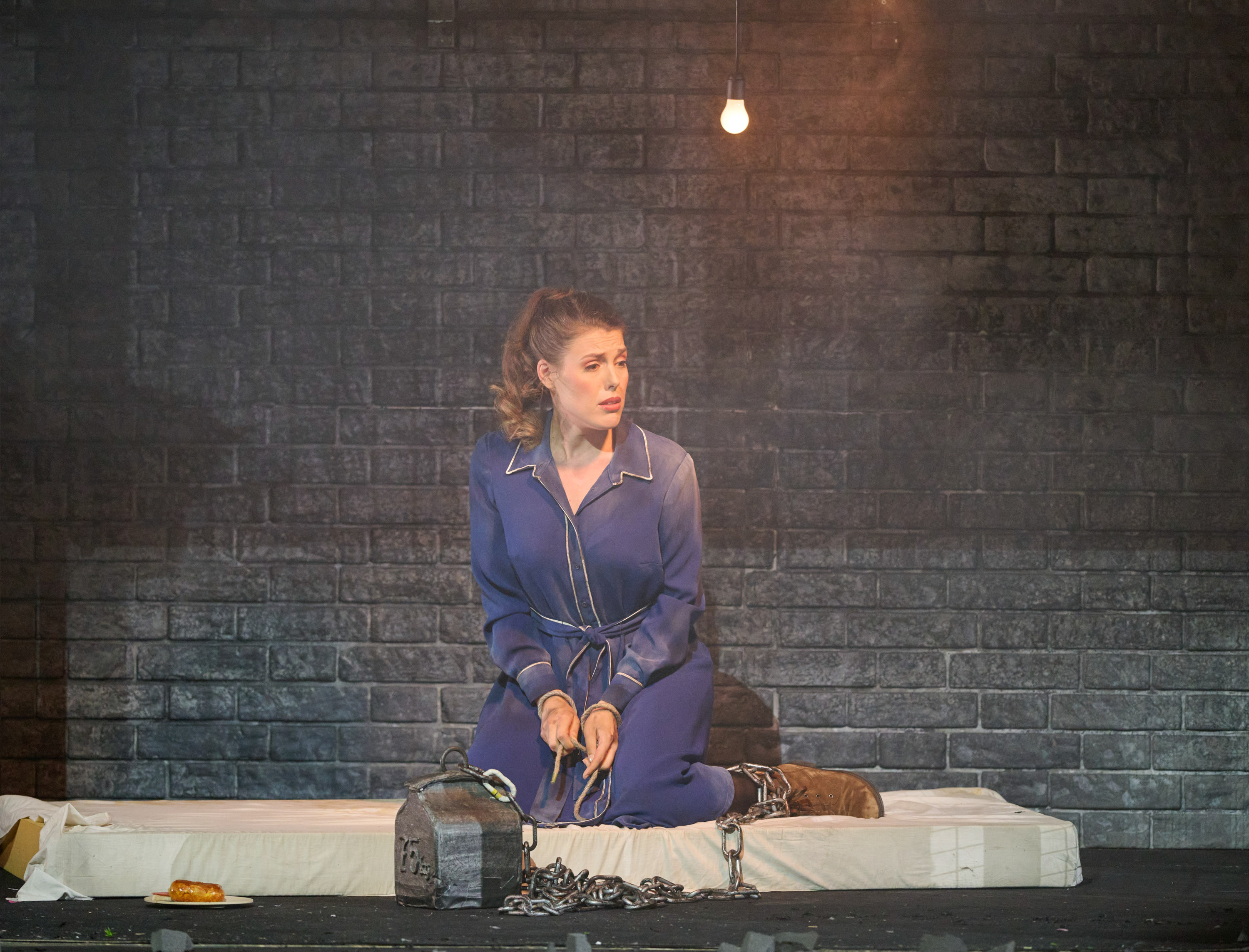 Eine Frau in einem schlichten blauen Kleid sitzt in Ketten gefesselt auf einer Matratze.