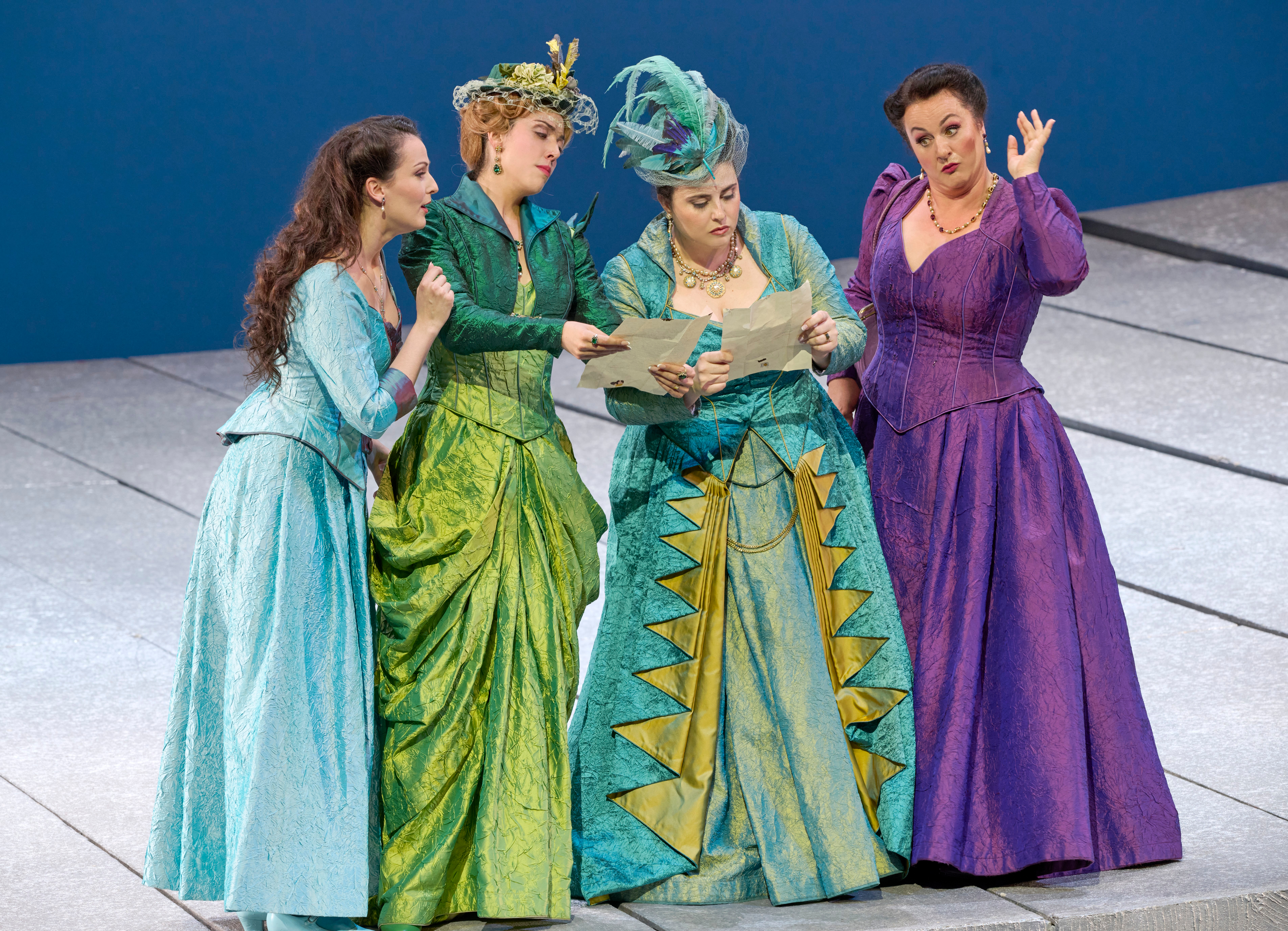 Vier Frauen in farbenfrohen Kostümen stehen auf der Bühne und schauen gemeinsam auf ein großes Blatt Papier.