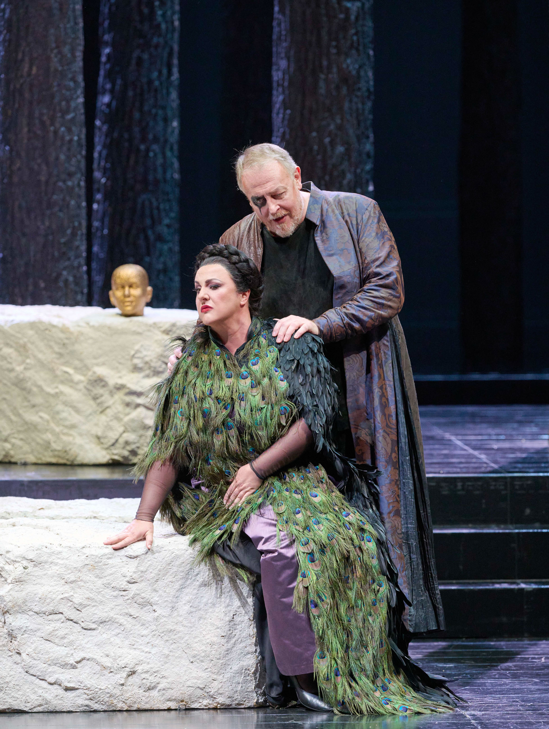 Ein Mann in langem Mantel steht hinter einer sitzenden Schauspielerin in Federumhang, während sie auf einer Bühne neben einer Statue eines Kopfes sind.