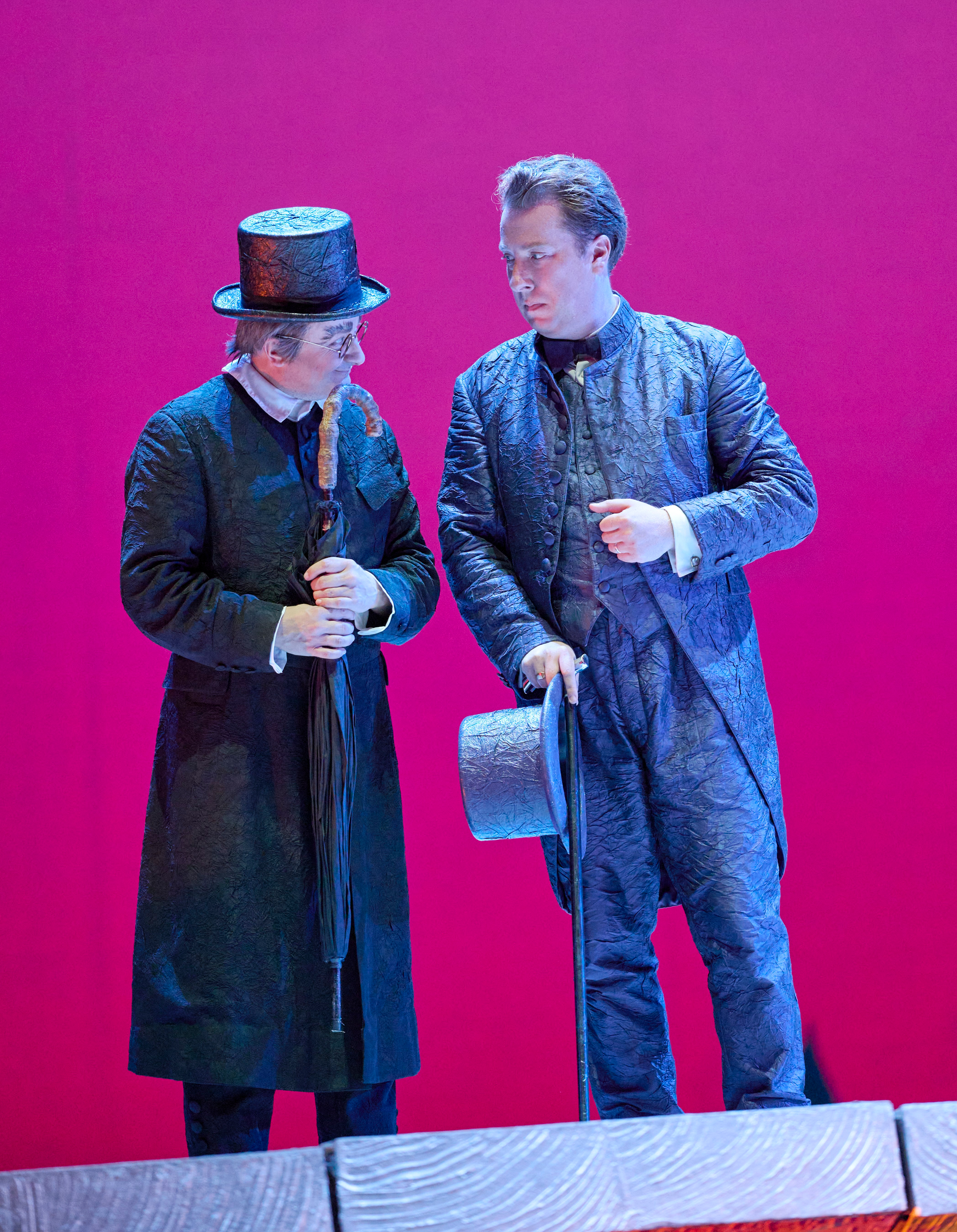 Zwei Männer in eleganter Kleidung stehen vor pinkem Hintergrund und unterhalten sich, einer hält einen Zylinder in der Hand.