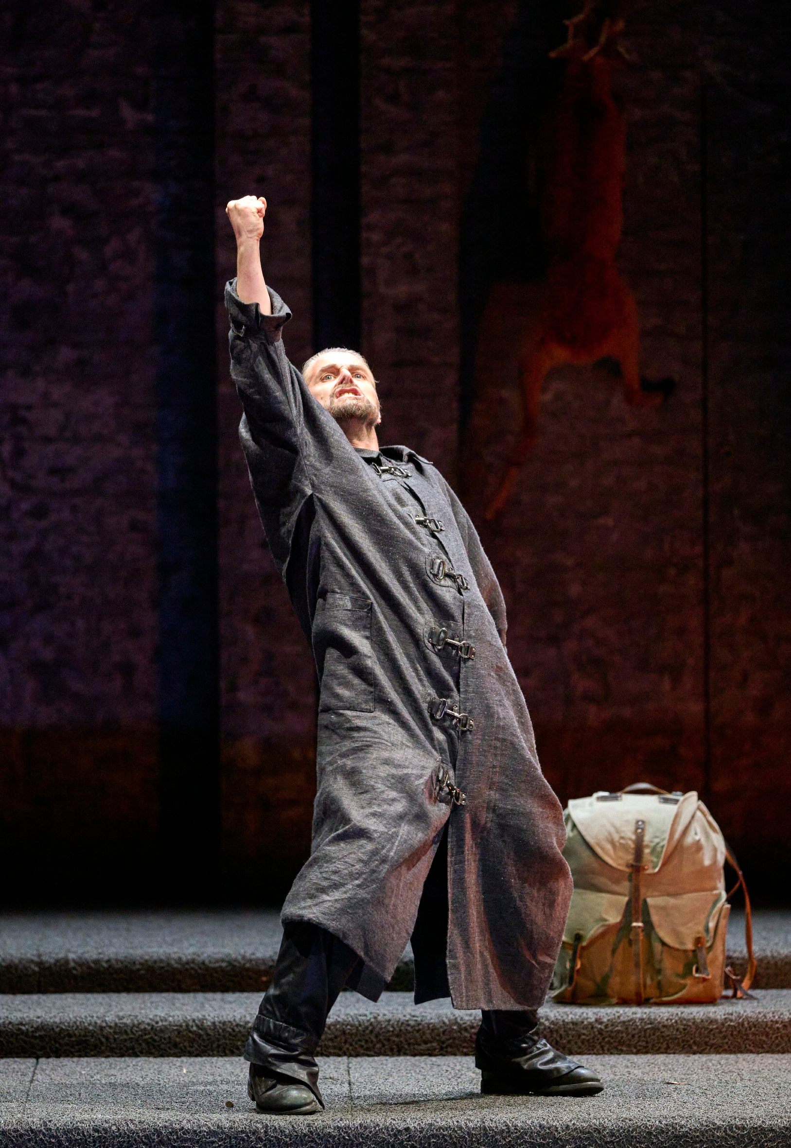 Ein Mann in einem grauen Overall streckt triumphierend die Faust in die Luft, im Hintergrund hängt ein Tierkadaver an der Wand.