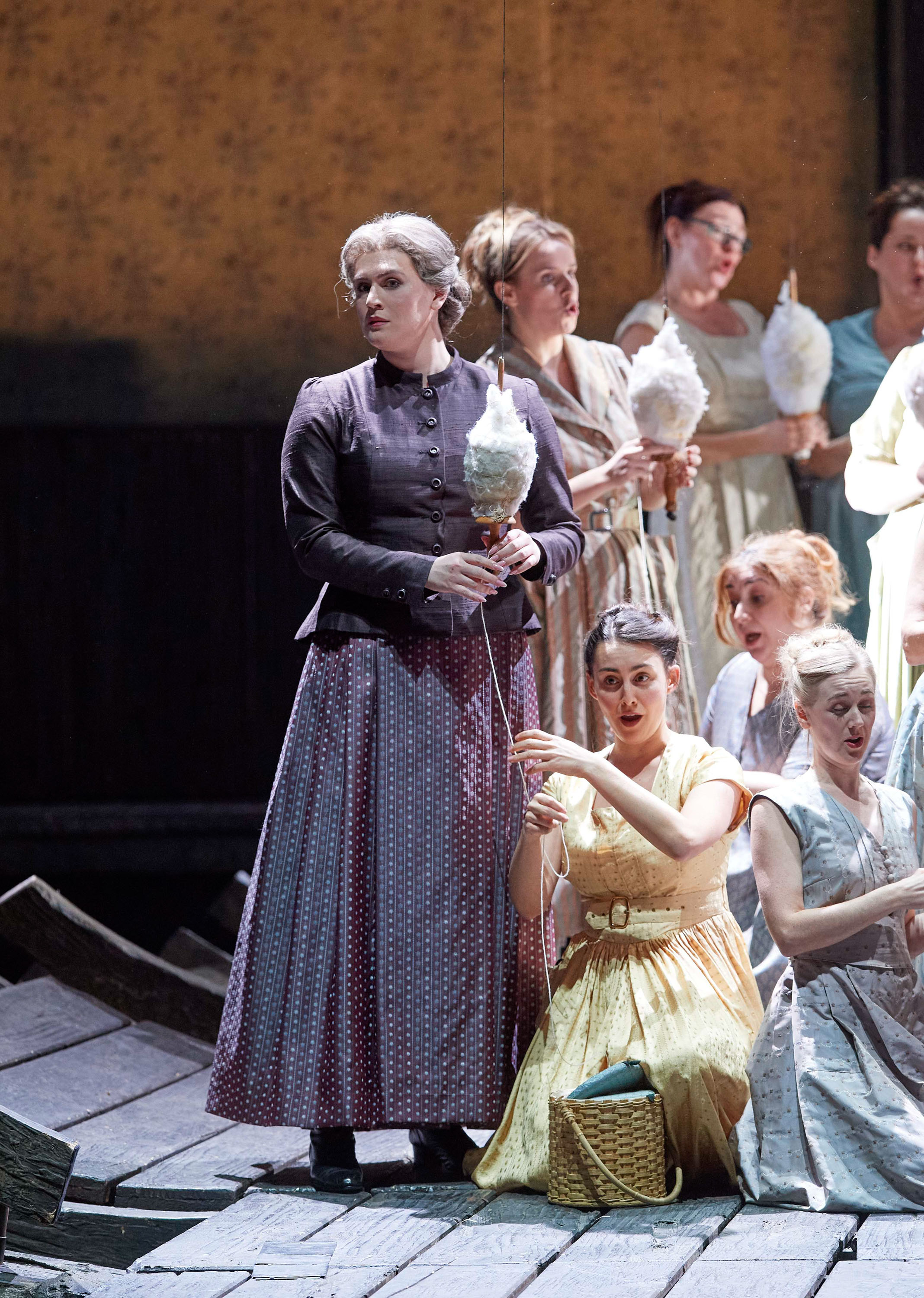 Eine ältere Frau in historischer Kleidung steht ernst mit einer weißen Feder in der Hand vor einer Gruppe junger Frauen in Pastellkleidern.