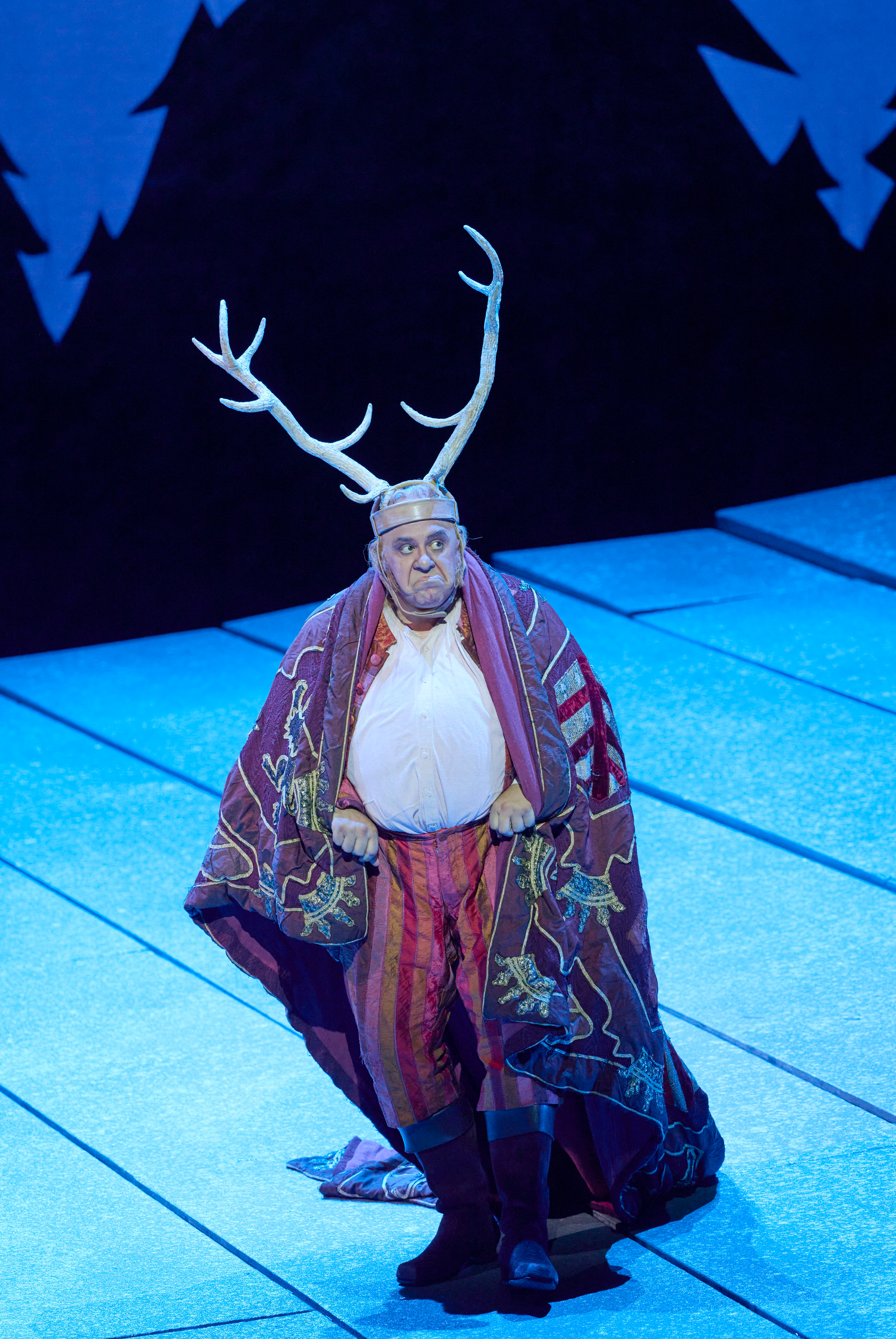 Eine Figur mit imposantem Geweih und buntem Umhang steht auf einer blau beleuchteten Bühne vor stilisierten Tannen.