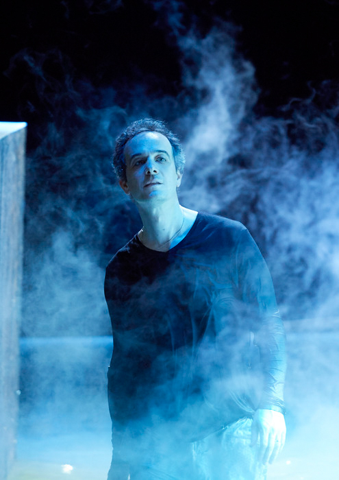 Ein Mann in dunkler Kleidung steht in geheimnisvollem, blauem Nebel und blickt versonnen nach oben.