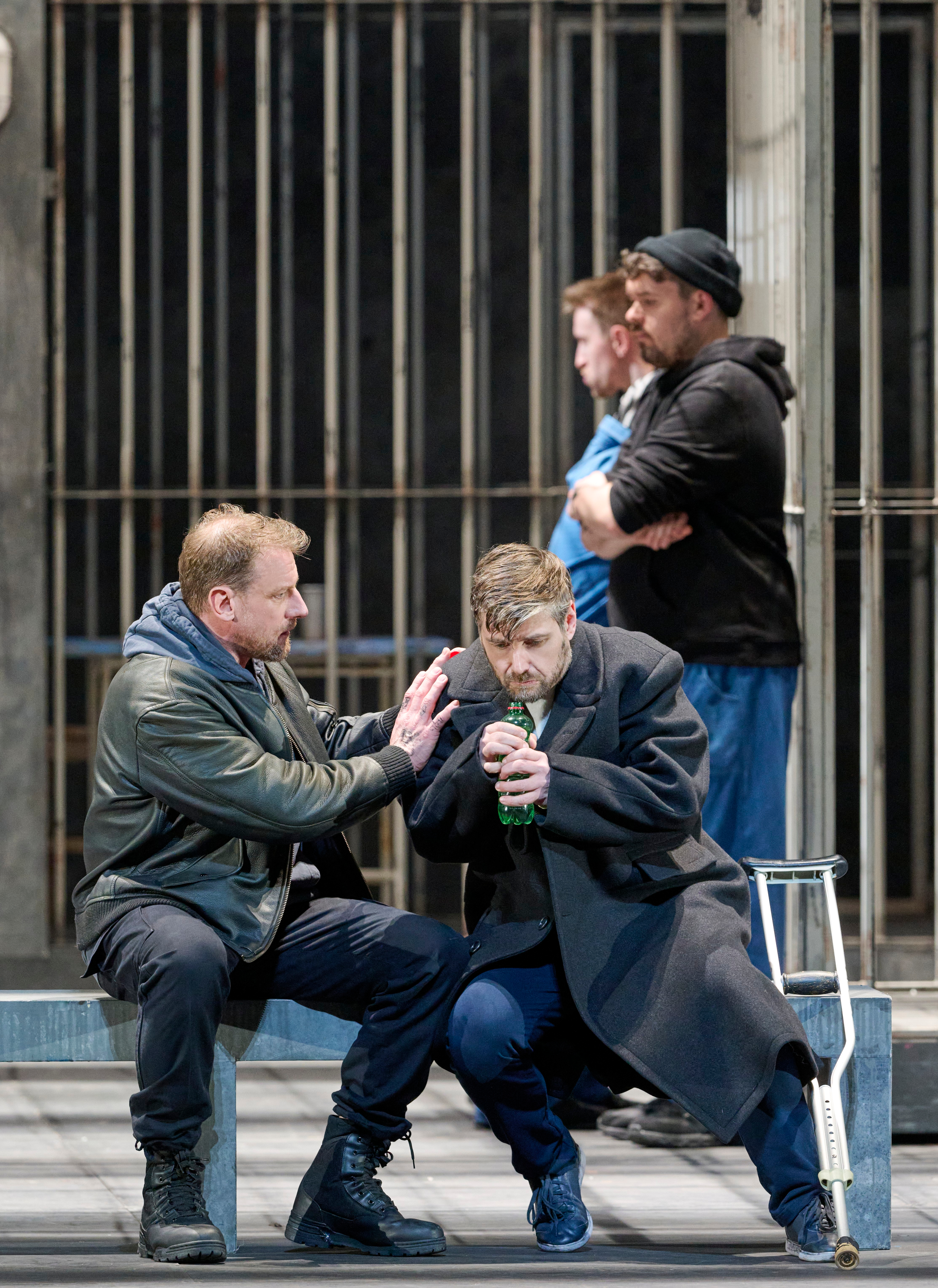 Zwei Männer sitzen vor einer Gitterwand, einer tröstet den anderen, während im Hintergrund zwei weitere Männer stehen und beobachten.