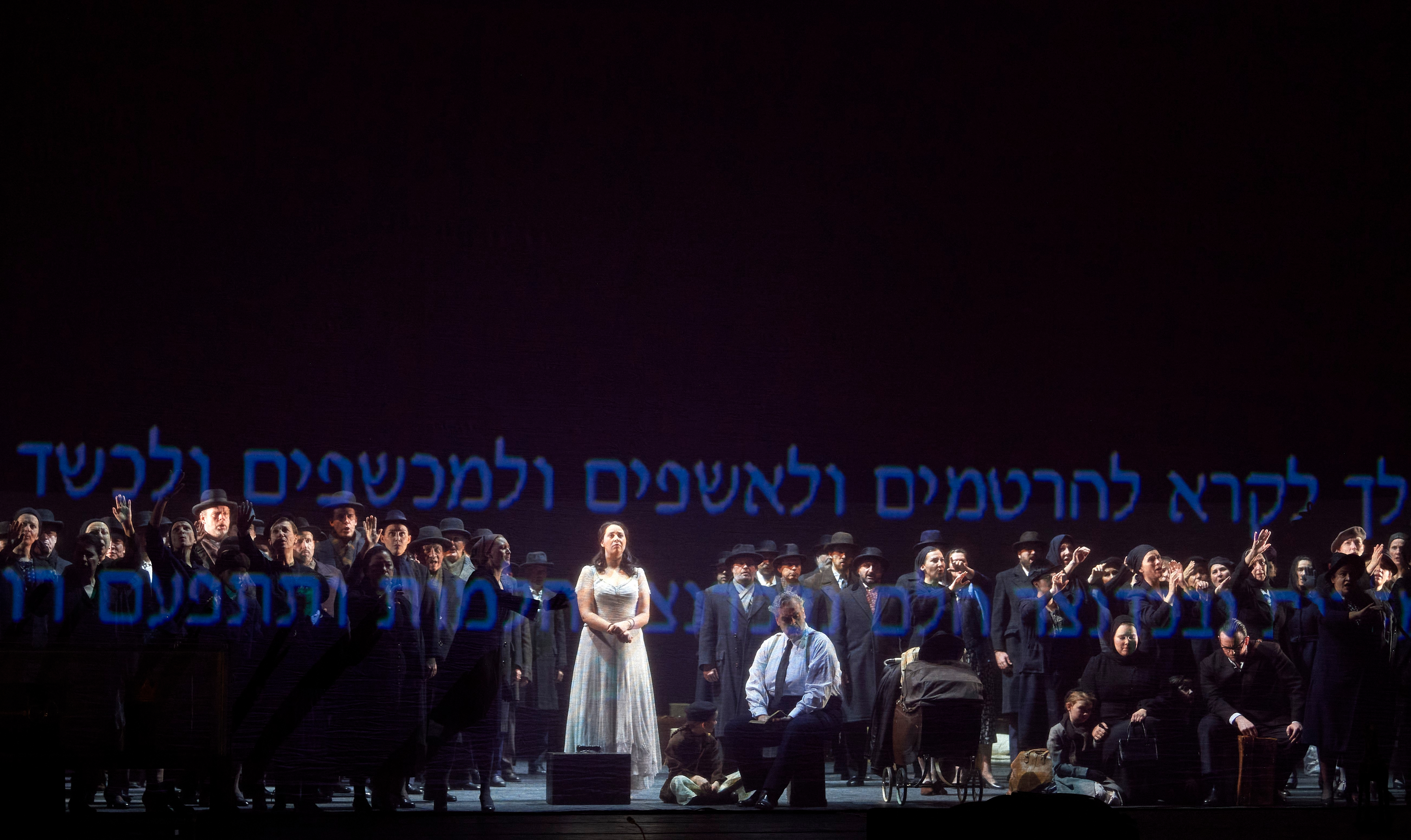 Eine Frau in hellem Kleid steht im Zentrum einer Gruppe dunkel gekleideter Menschen auf einer Bühne mit hebräischer Schriftprojektion.