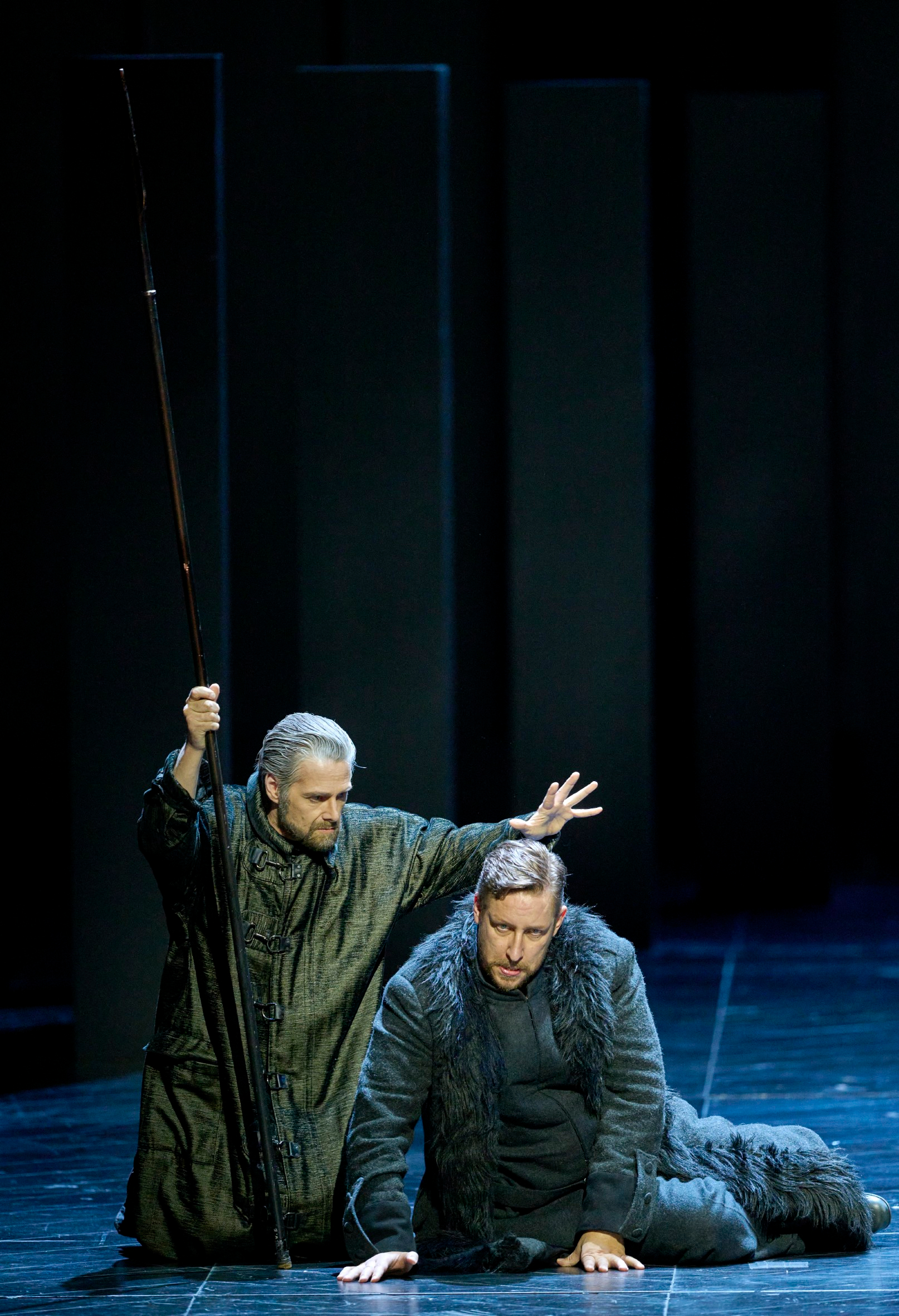 Zwei Männer in dunklen Gewändern befinden sich auf einer düsteren Bühne; einer kniet mit gesenktem Blick, während der andere eine segnende oder beschwörende Geste über ihn macht.