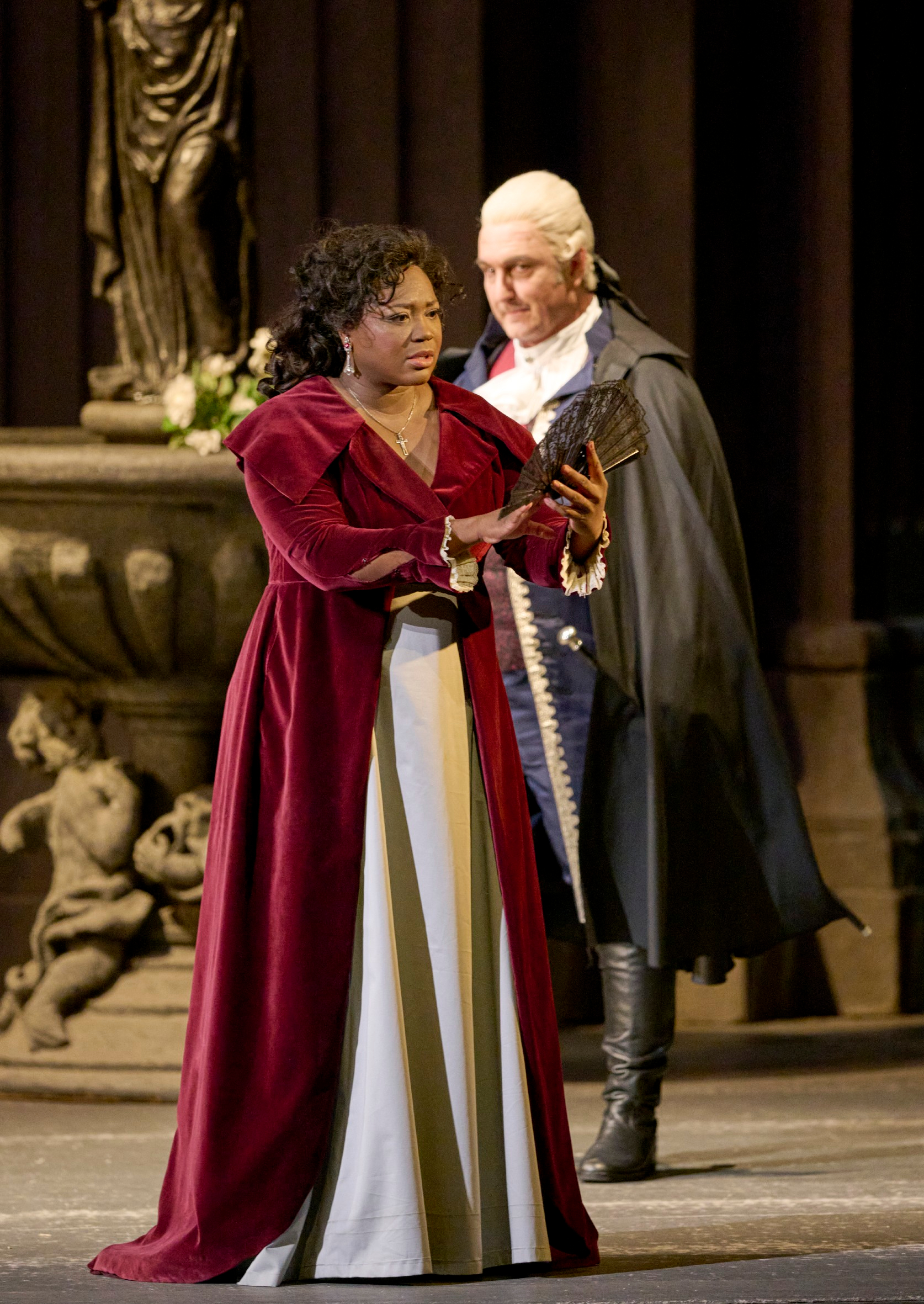 Eine Frau in einem roten, eleganten Kleid hält einen Fächer und spricht mit einem Mann in einem dunkelblauen Mantel vor einer Statue.