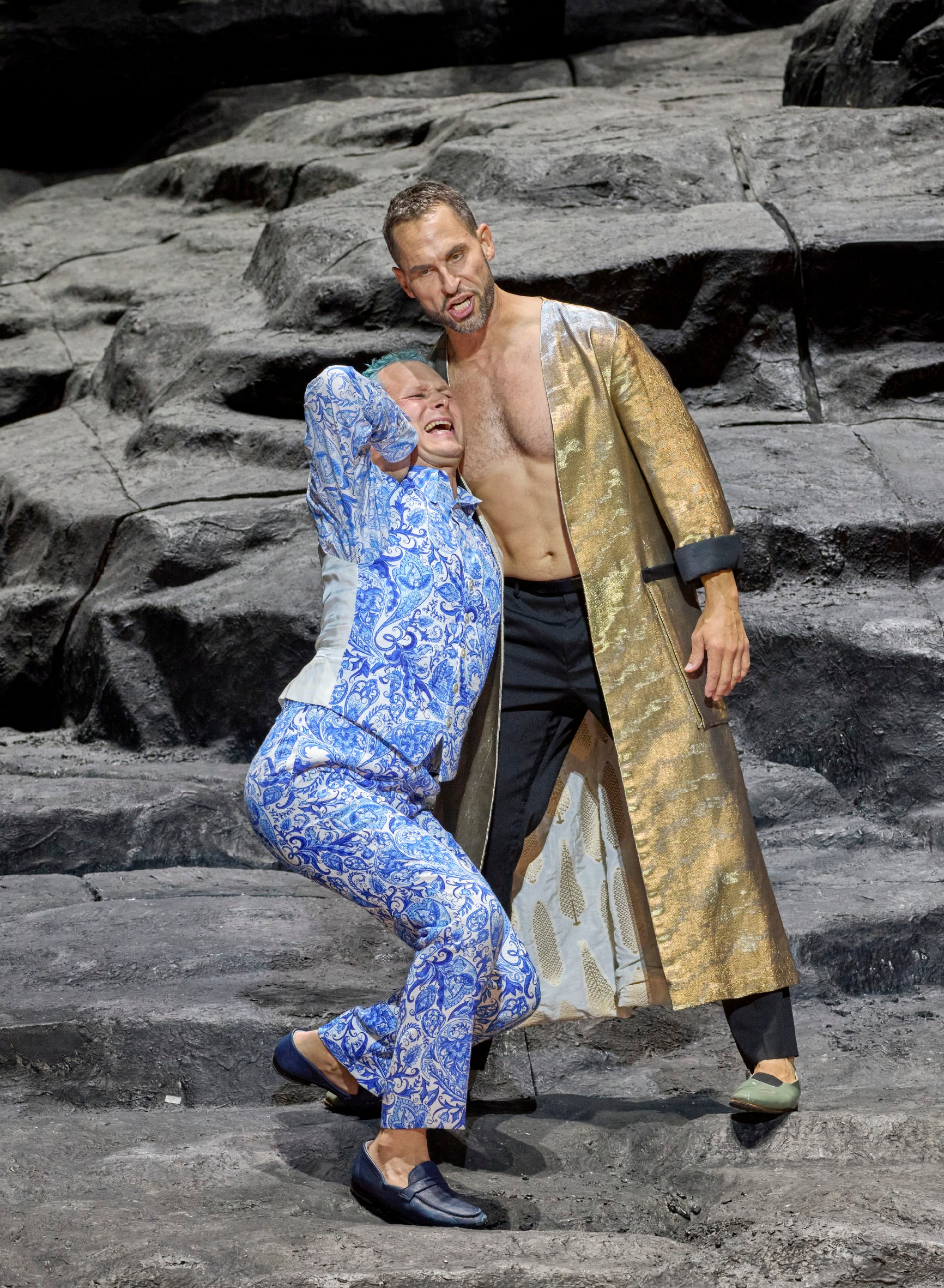 Ein Mann im blauen Anzug schreit während ein anderer Mann im goldenen Mantel ihn am Rücken packt.