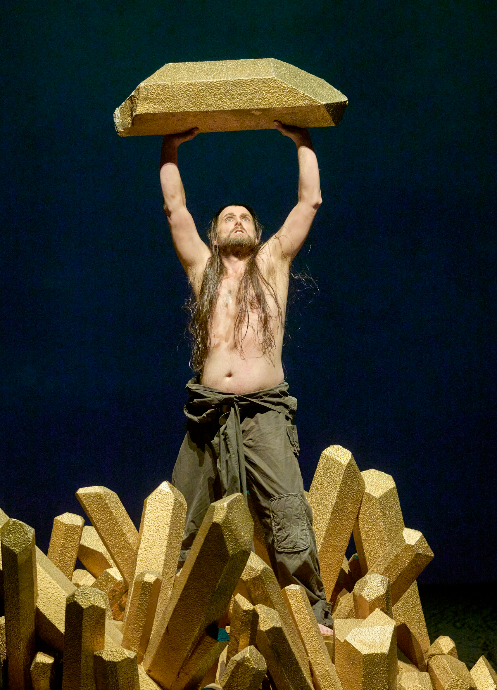 Ein barfüßiger Mann mit langem Haar und nacktem Oberkörper hebt feierlich einen goldenen Block über den Kopf.