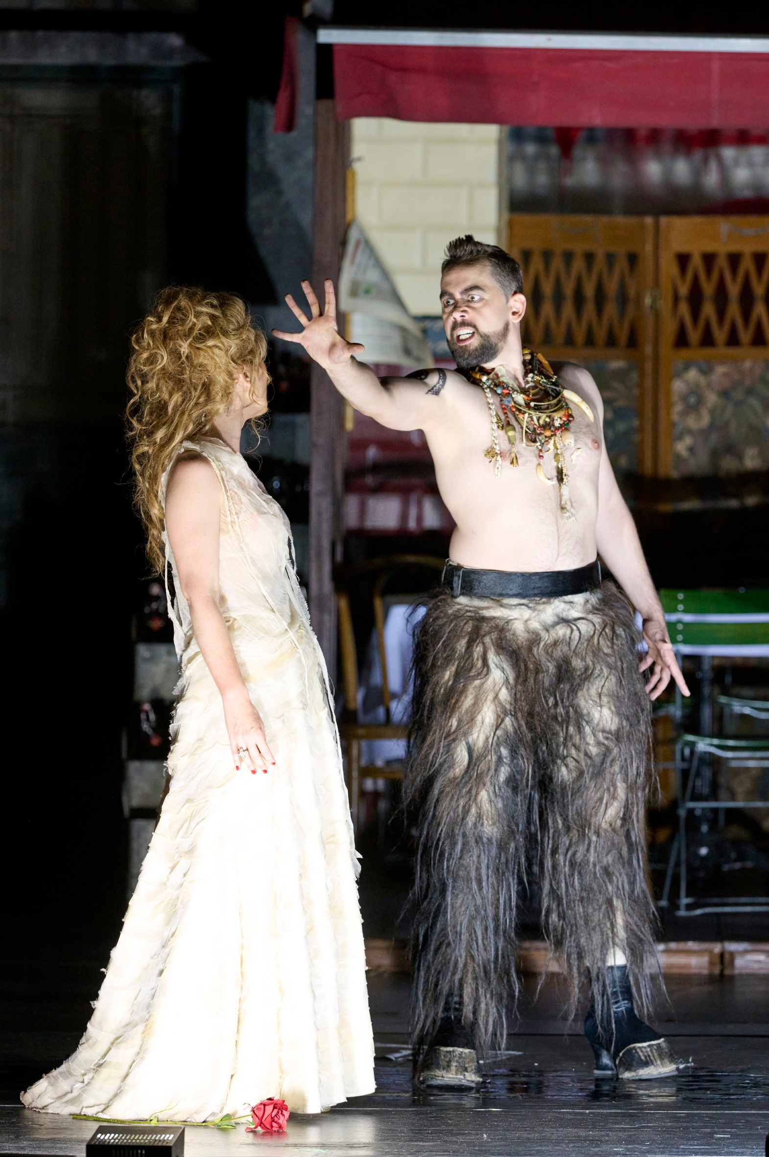 Ein Mann mit nacktem Oberkörper und wildem Kostüm streckt dramatisch die Hand aus, während eine Frau im weißen Kleid ihn ruhig ansieht.