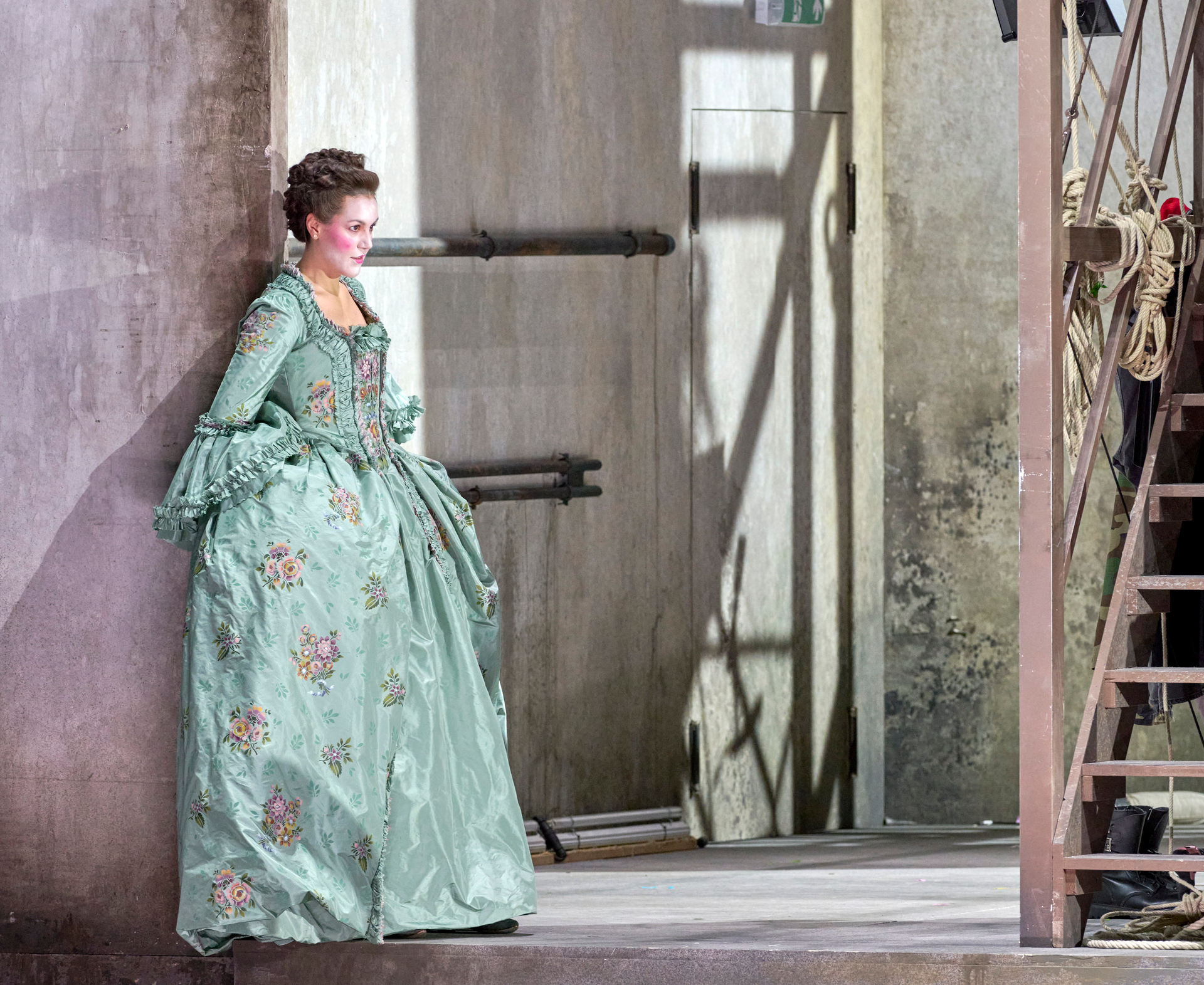 Eine Frau in einem opulenten, hellgrünen barocken Kleid mit Blumenmuster lehnt an einer Wand und blickt mit leichtem Lächeln zur Seite.
