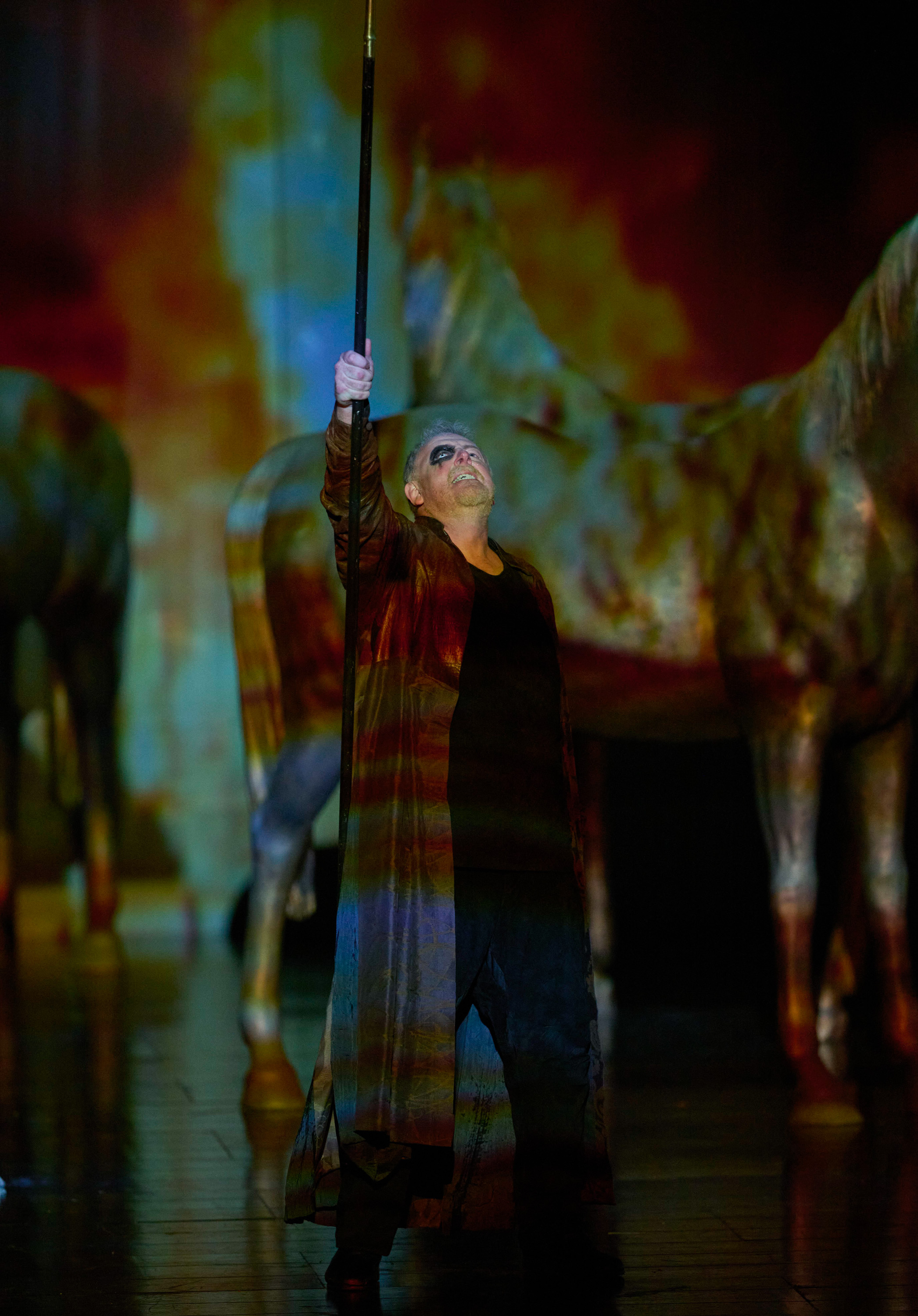 Eine Figur in langem Mantel hält eine Stange in die Luft, während im Hintergrund riesige Pferde zu sehen sind.