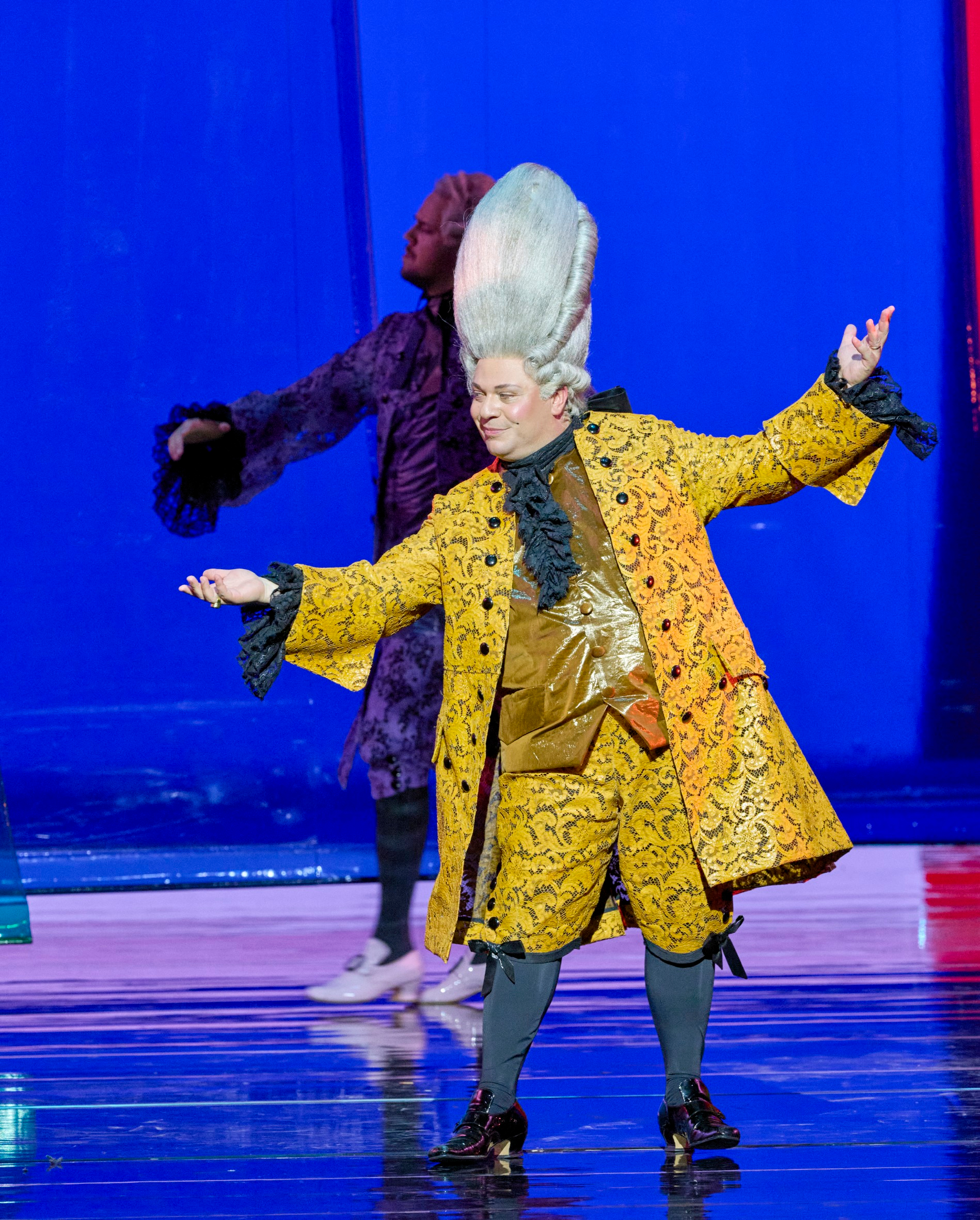 Ein Darsteller in prunkvollem gelbem Rokoko-Kostüm und hoher weißer Perücke posiert theatralisch auf der Bühne vor blauem Hintergrund.