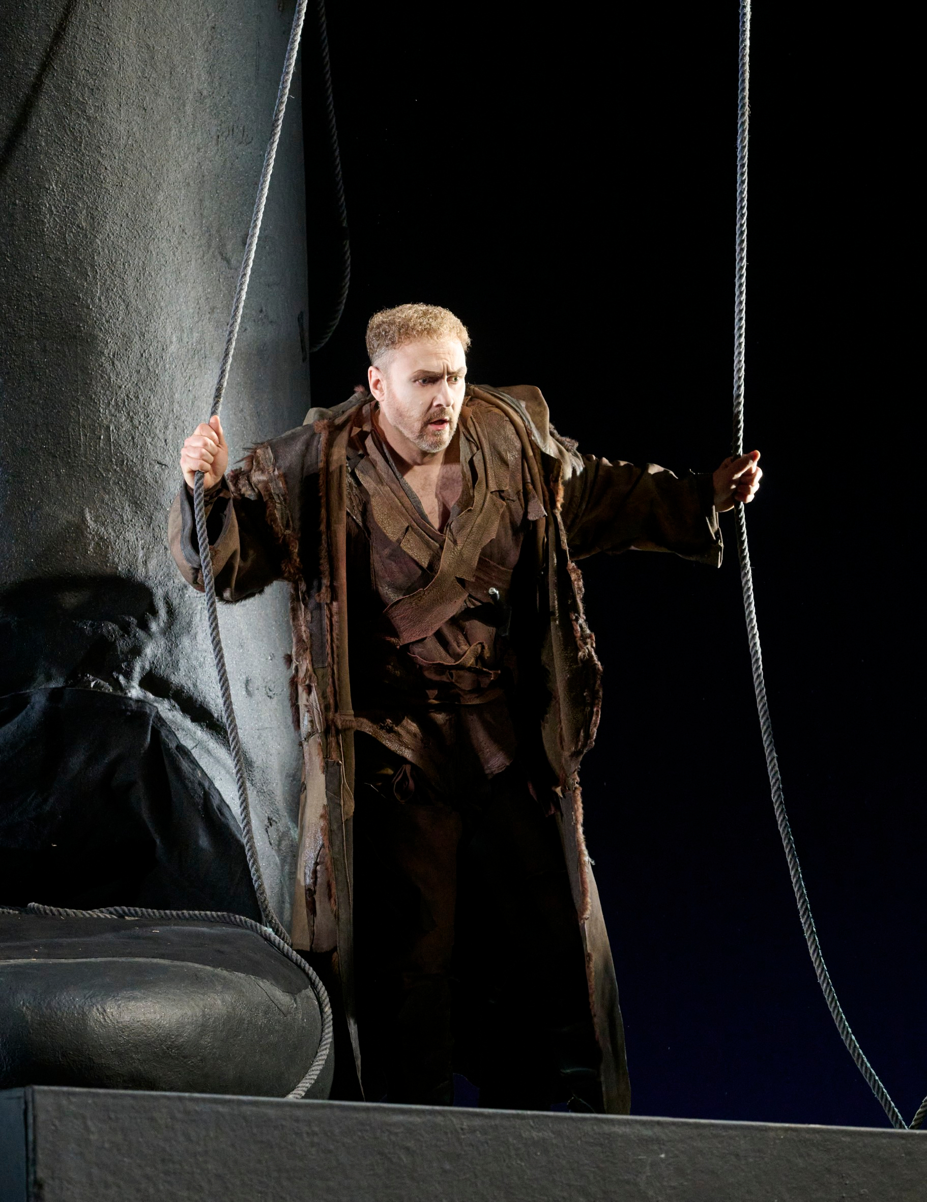 Ein Mann in braunem Bühnenkostüm hält zwei Seile und blickt ernsthaft nach vorn.