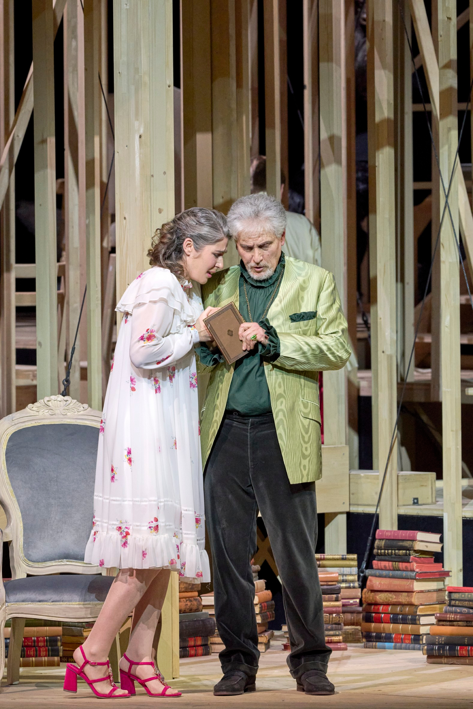 Eine Frau im geblümten Nachthemd und ein Mann im grünen Jackett betrachten gemeinsam ein Buch vor einer hölzernen Bühnenkonstruktion.