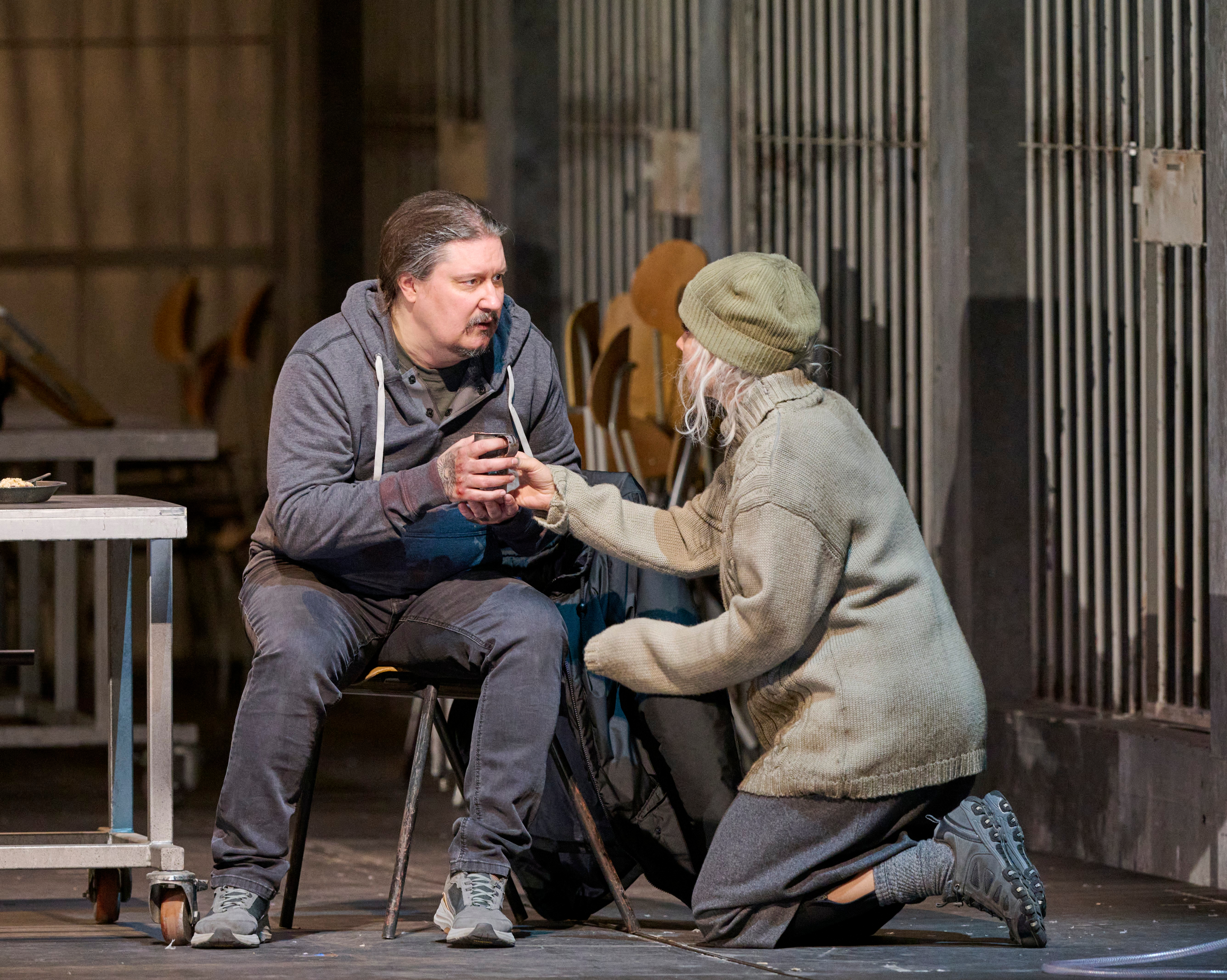 Ein Mann sitzt mit einem Becher in der Hand, während eine Frau in warmer Kleidung kniend seine Hände berührt.