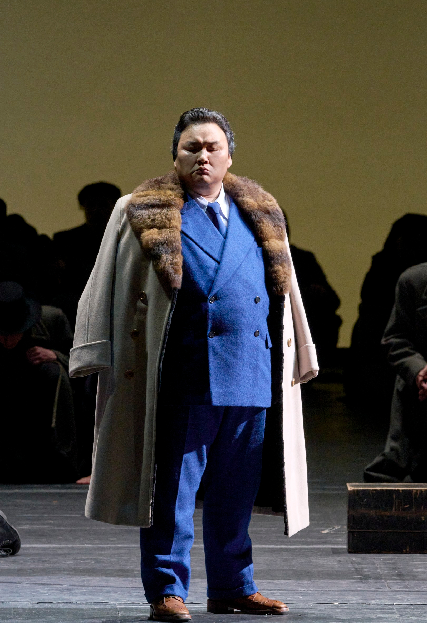 Ein Mann im königsblauen Anzug und cremefarbenem Mantel mit Pelzkragen steht selbstbewusst vor einer Gruppe dunkler Silhouetten.