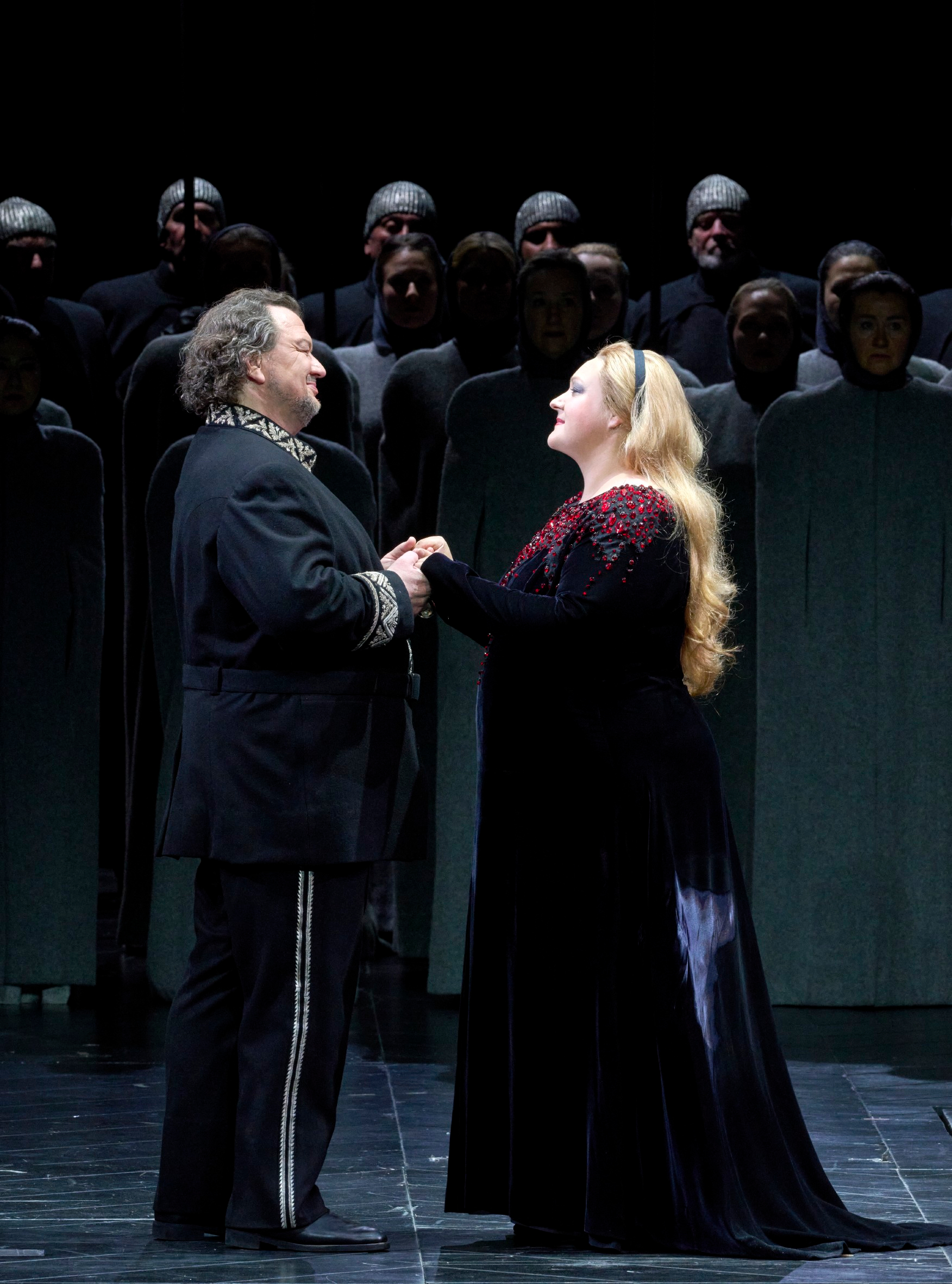 Ein Mann und eine Frau halten sich an den Händen und singen ein Duett, während ein Chor in dunkler Kleidung im Hintergrund steht.