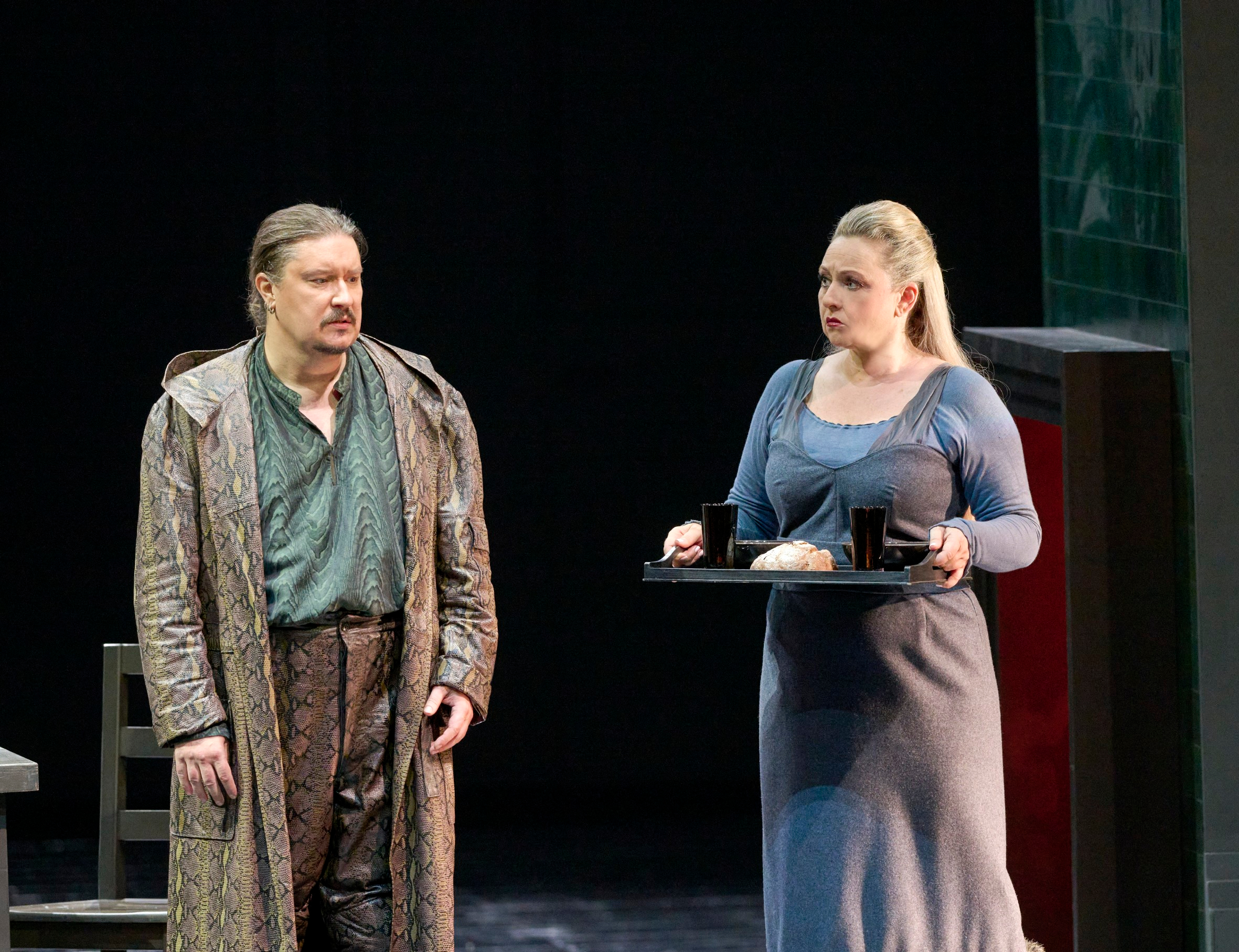 Ein Mann in gemustertem Mantel steht neben einer Frau in grauem Kleid, die ein Tablett mit Tassen und einem Brötchen hält.